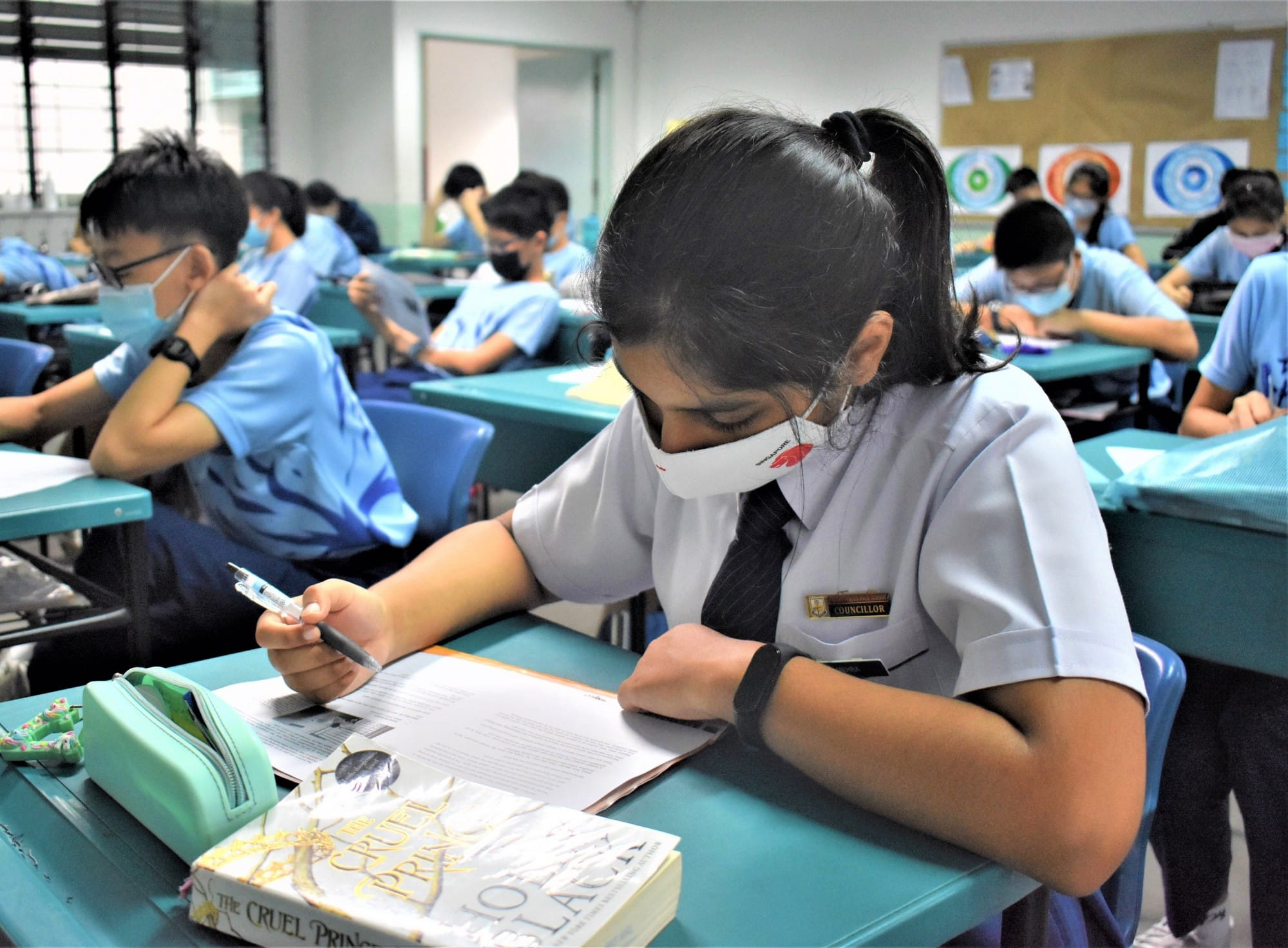 Students in school uniforms and face masks sit at teal desks writing, with a girl in the foreground reading "The Cruel Prince."