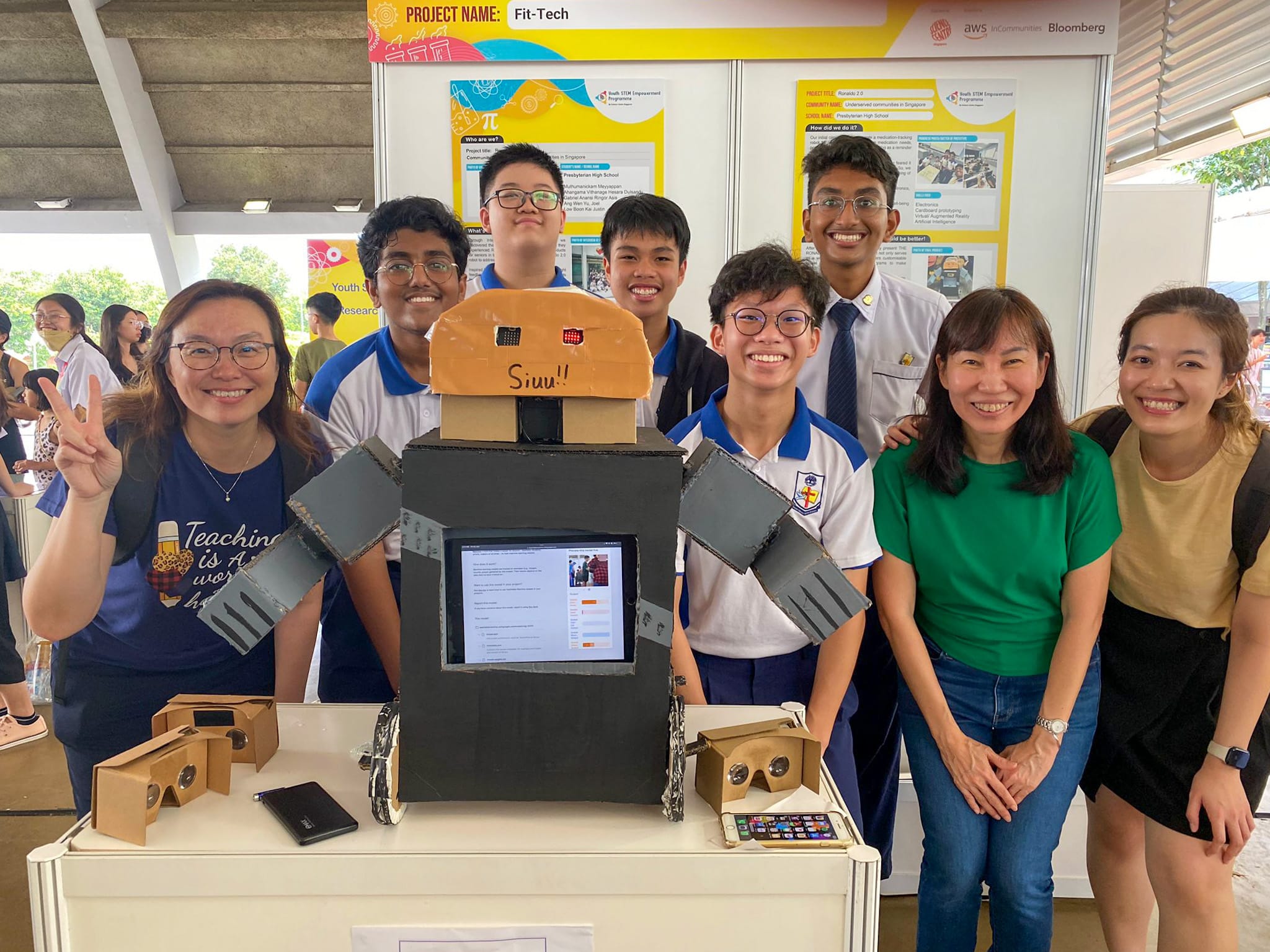 Seven smiling people pose with a cardboard robot labeled Siuu at a Fit-Tech STEM project fair booth.