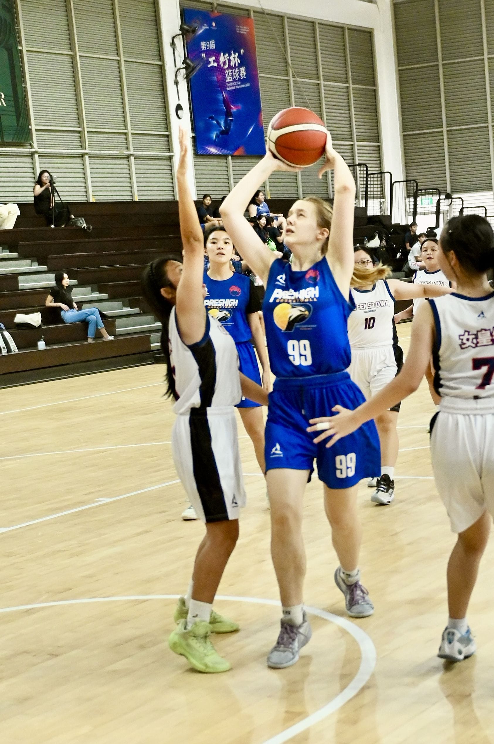 Player number 99 in blue shoots a basketball over a defender during an indoor women's basketball game.