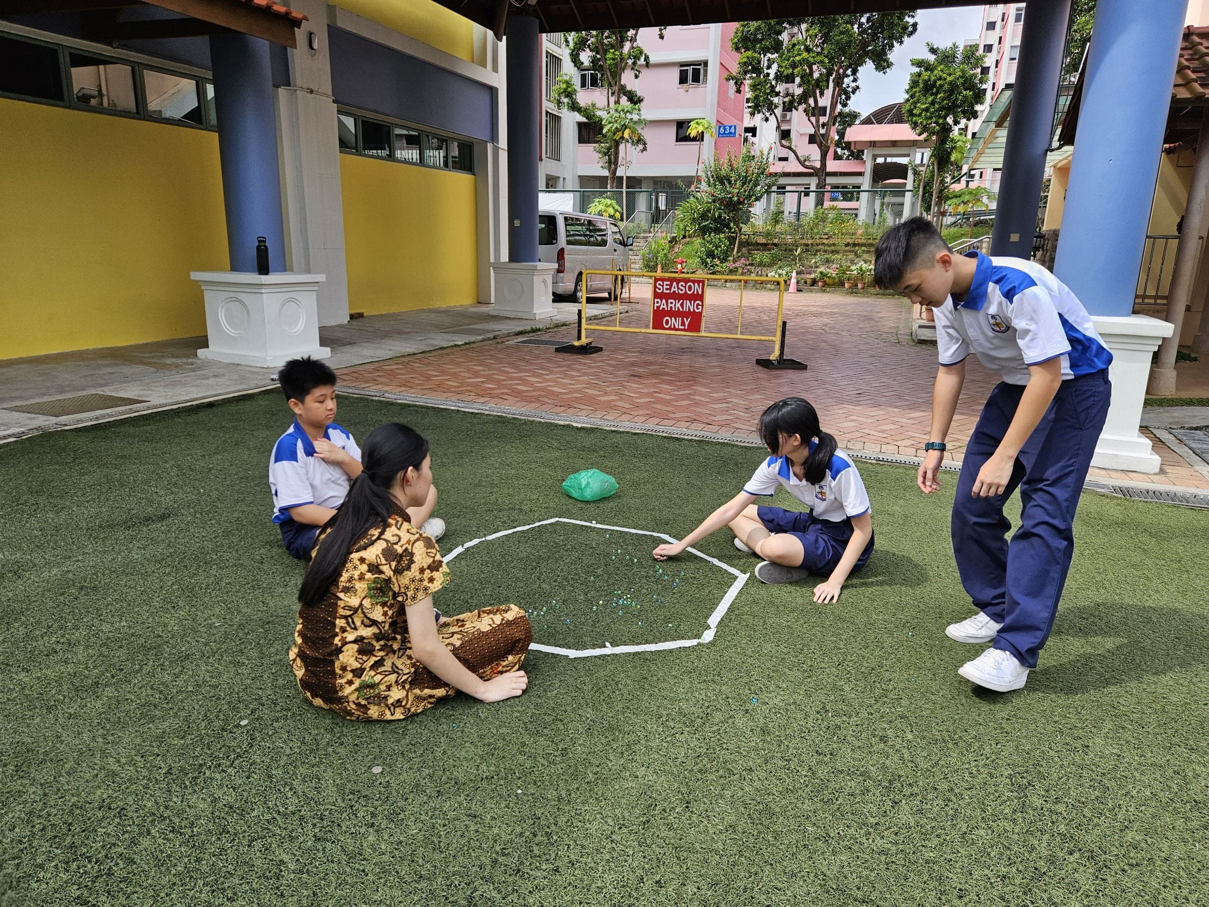 Four children in school uniforms play a marble game inside a taped circle on artificial grass outdoors.