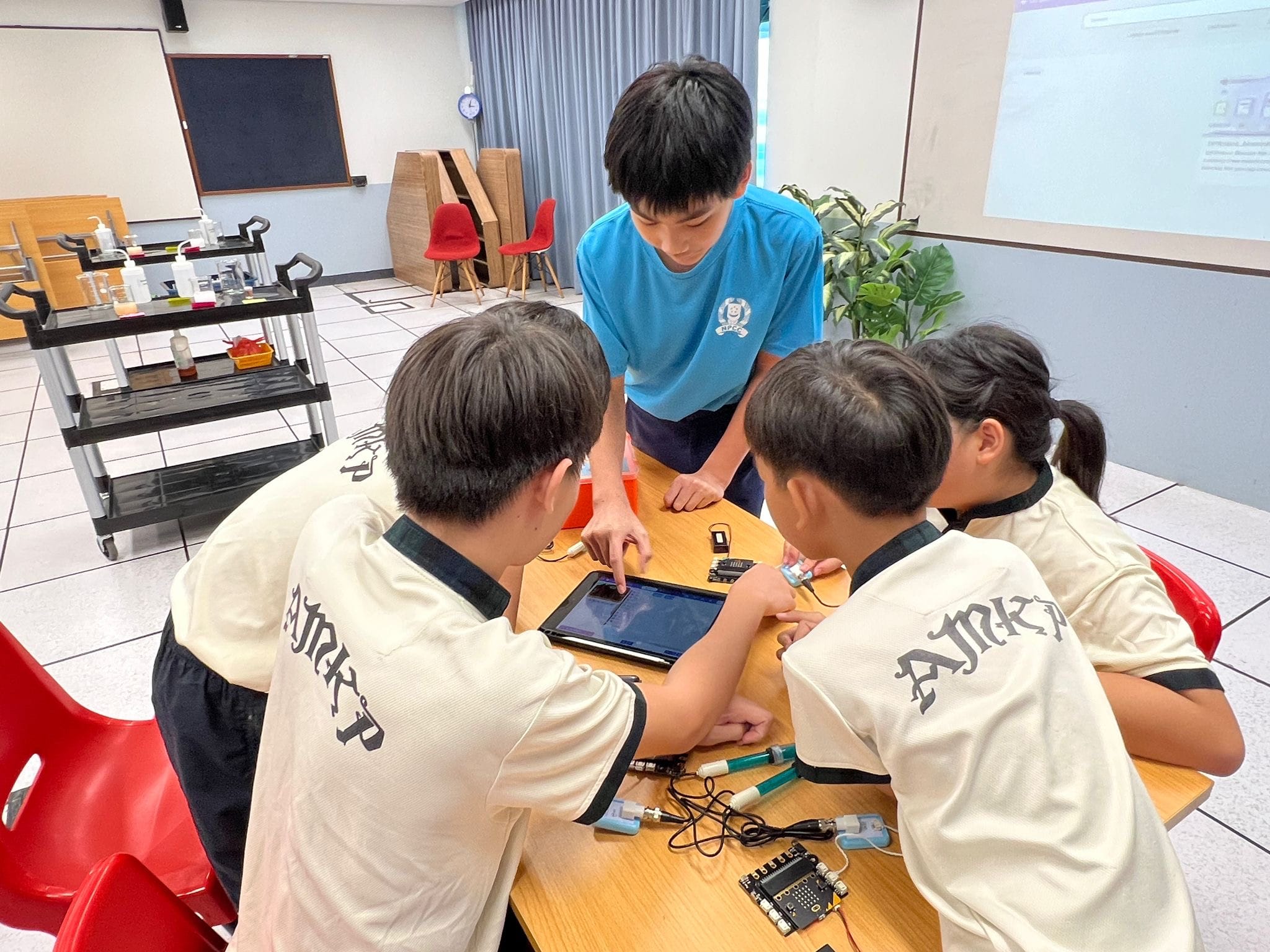 Four children gather around a table with a tablet and electronic circuit components in a classroom setting.