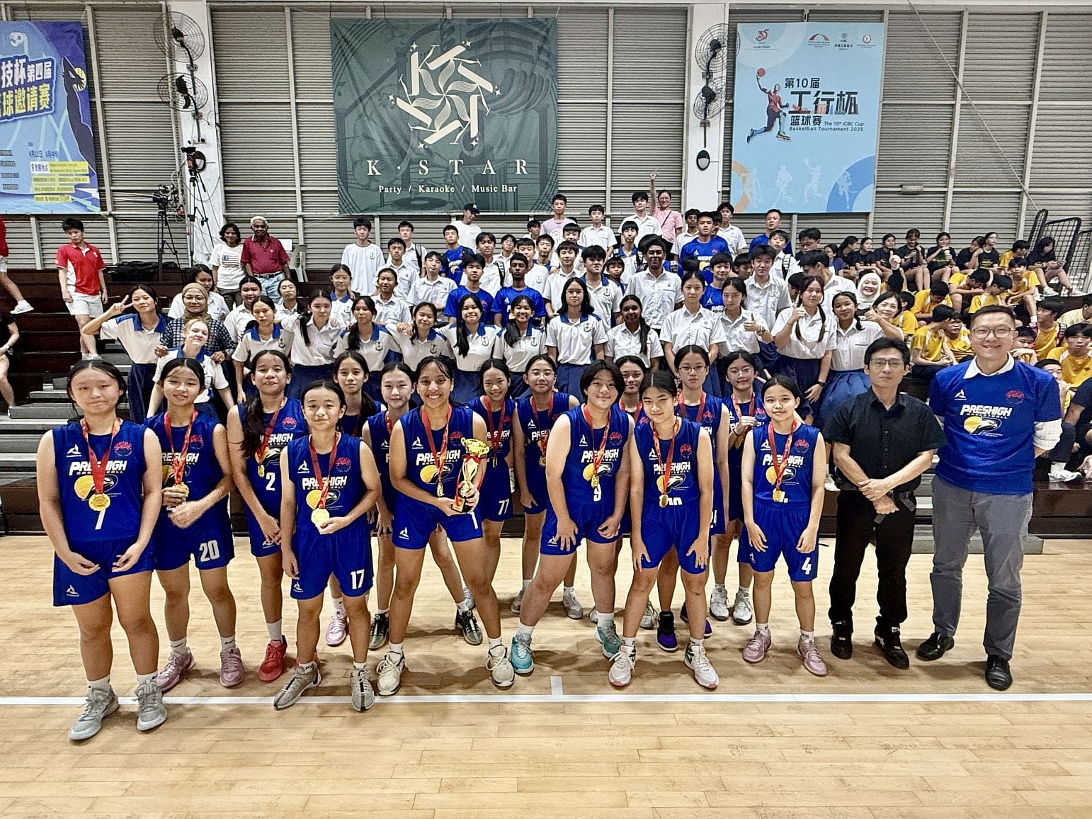 Girls basketball team in blue jerseys holding gold medals and a trophy, posing in a gym with a crowd behind them.