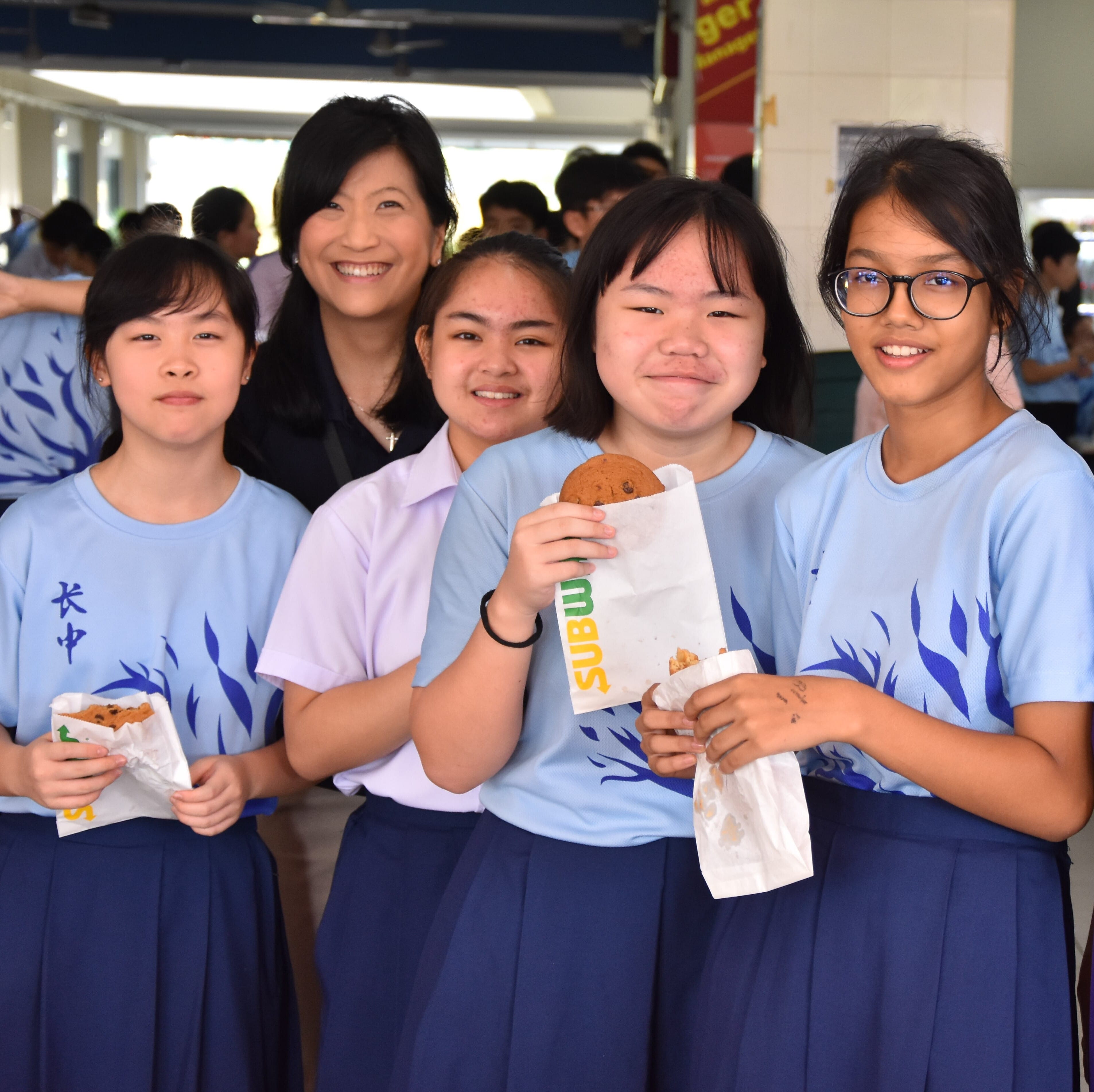 Four girls in blue school uniforms and an adult woman smile together, holding Subway cookie bags indoors.