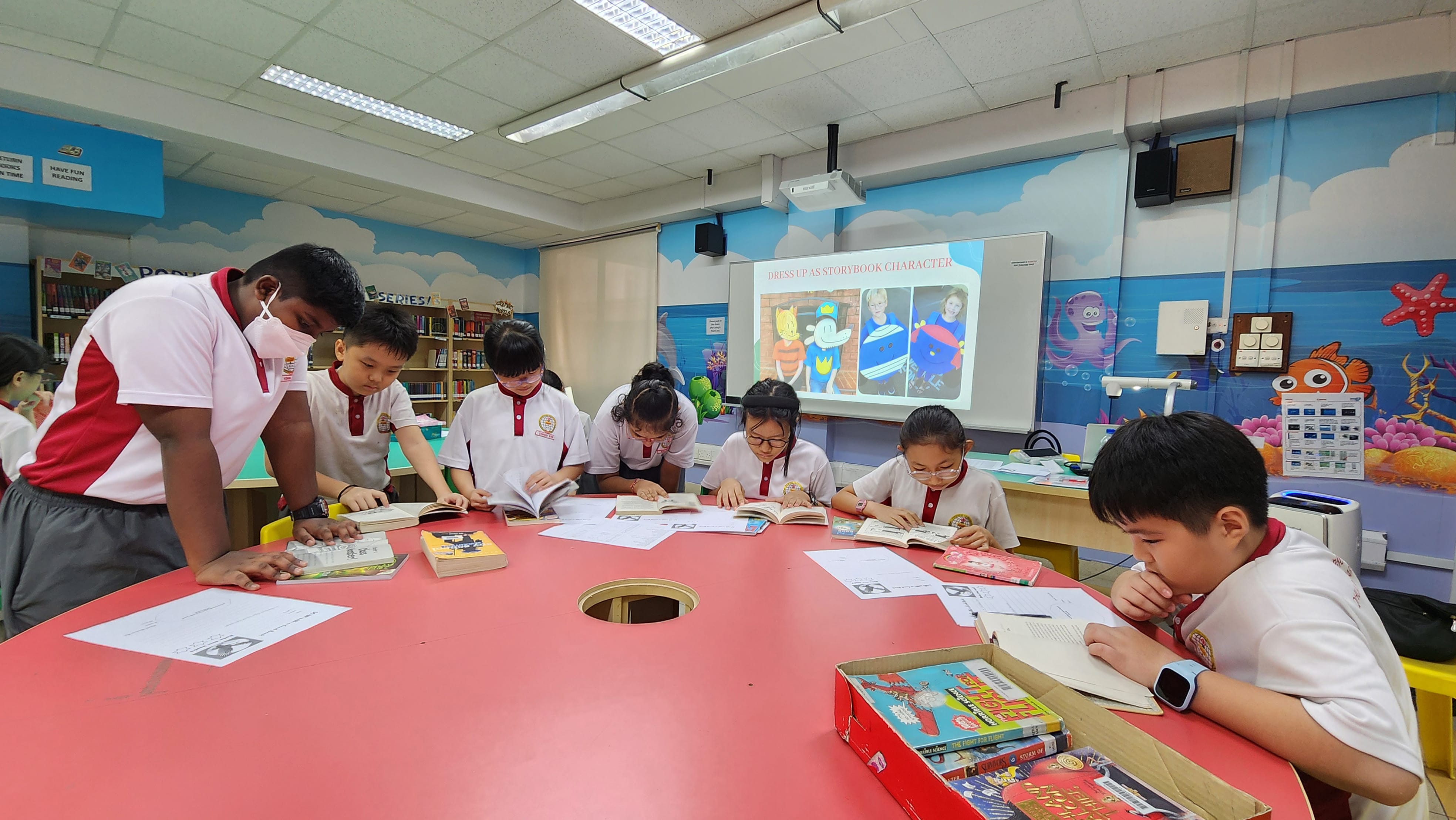 Several primary school children in uniforms read books at a large round red table in a colorful library.