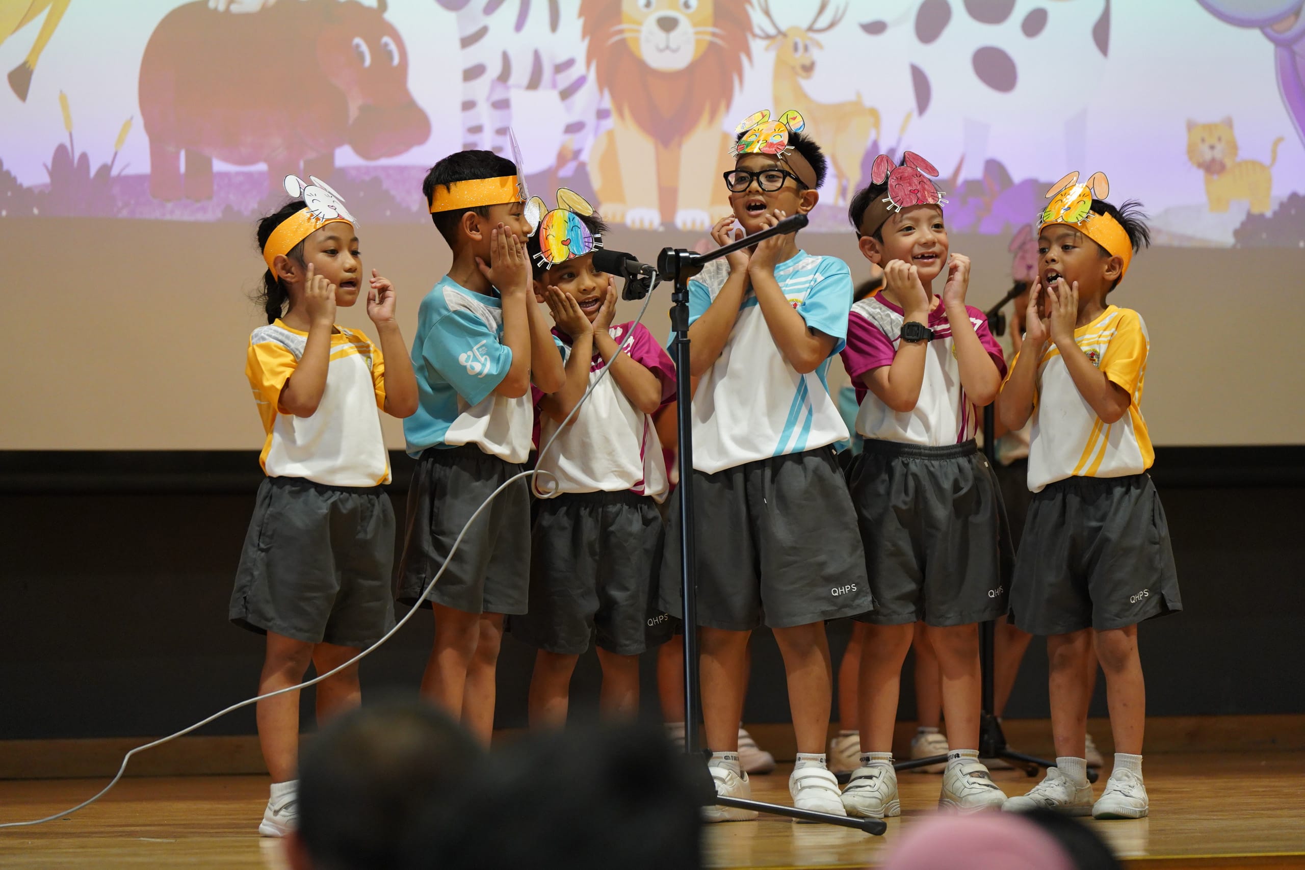 Students wearing animal headgear performing on stage singing to microphones on stands