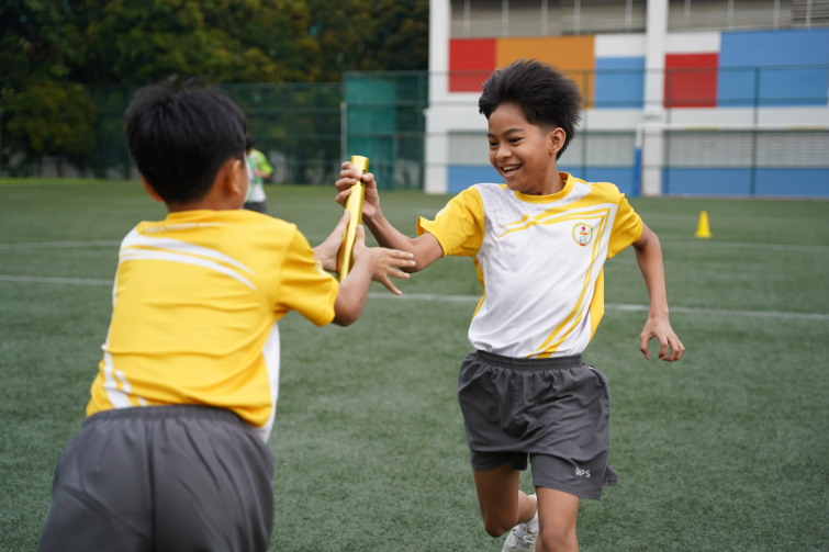 Boy in a yellow house PE attire passing the baton to another boy in an open field