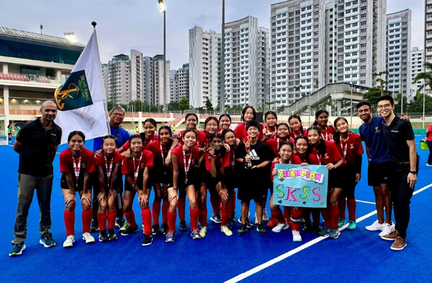 Girls hockey team in red uniforms posing with medals, a flag, and a Champion sign on a blue field.