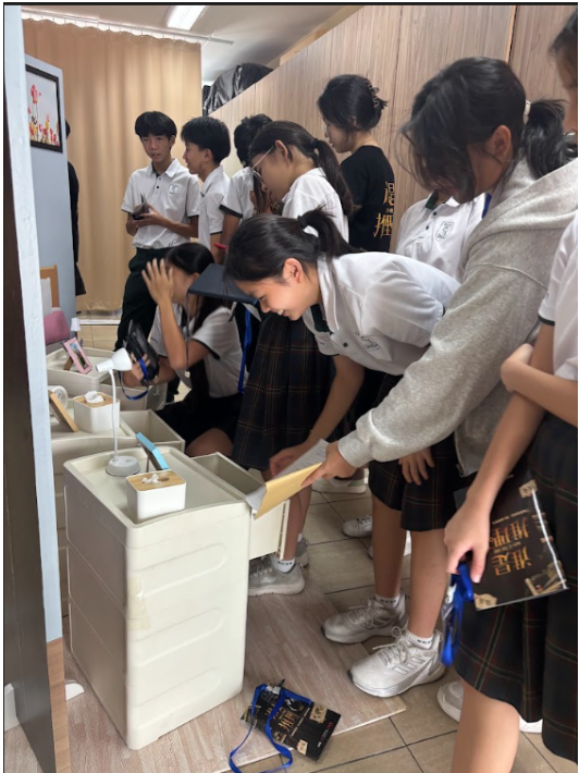 A group of students in school uniforms gathers around a display cabinet in an indoor room. Several students are examining items stored in drawers and compartments, while others observe closely. The display includes small objects and informational materials, and some students are holding booklets on lanyards. Wooden cabinets and a curtain are visible in the background.