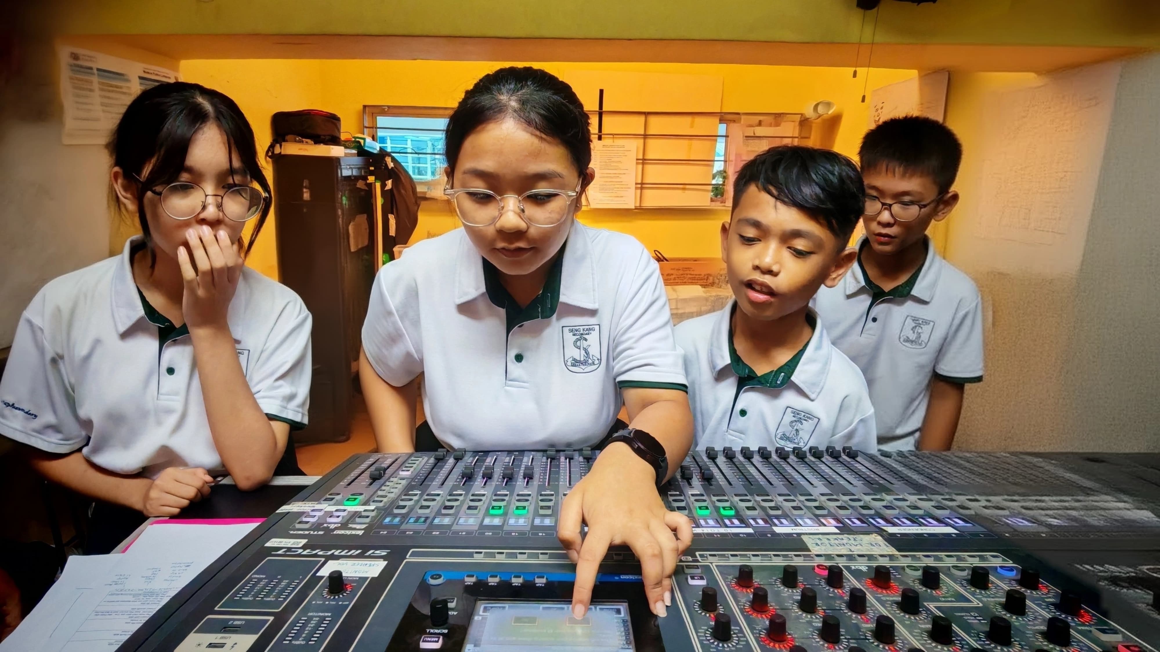 Four school students in white uniforms gathered around a mixing console, with one girl operating the controls. She is teaching her juniors how the mixer can be operated.