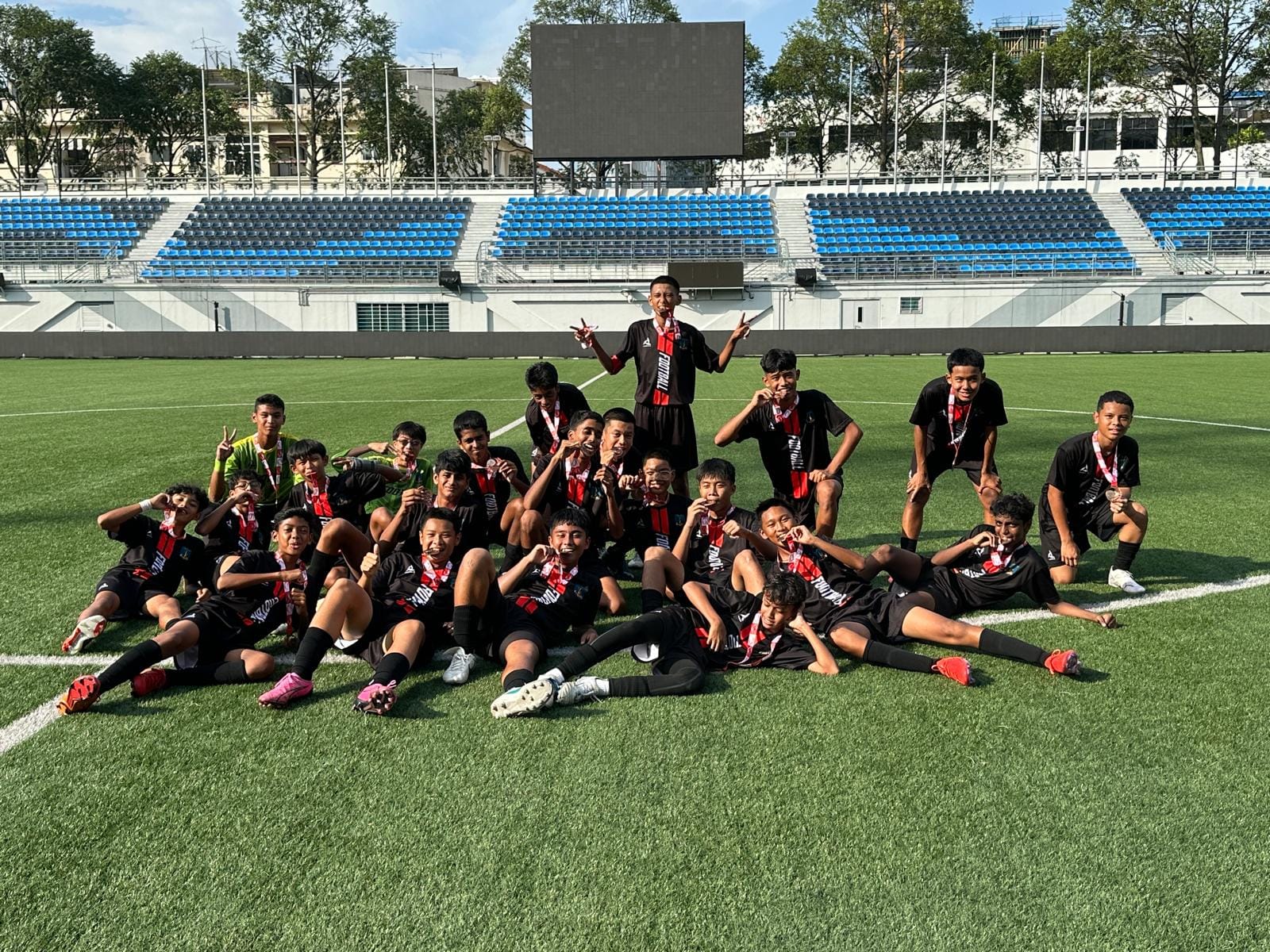 A youth soccer team in black uniforms poses with medals on a green field inside a stadium with blue seats.