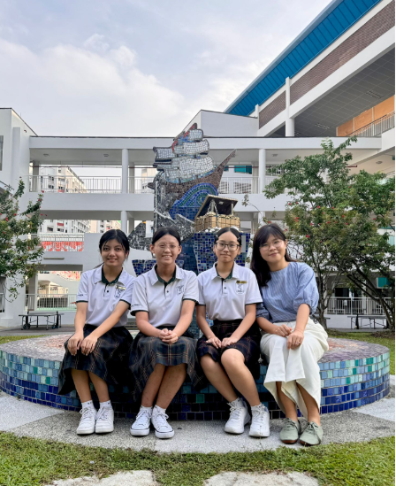 Four girls in school uniforms sit on a mosaic bench in front of a large mosaic sculpture at a school courtyard.