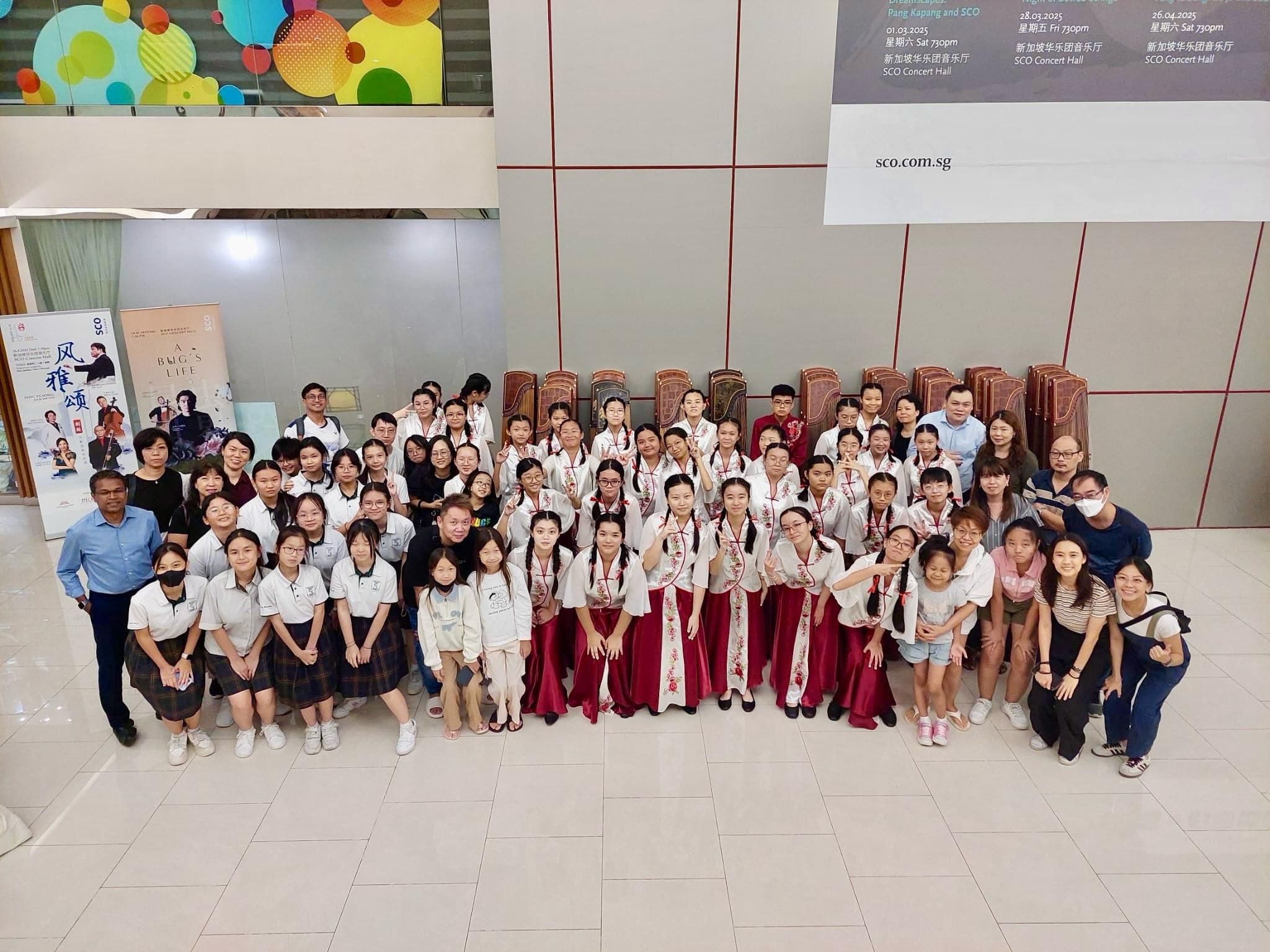 Large group of students and adults posing indoors, some wearing white and red traditional Chinese outfits, near SCO concert hall banners.