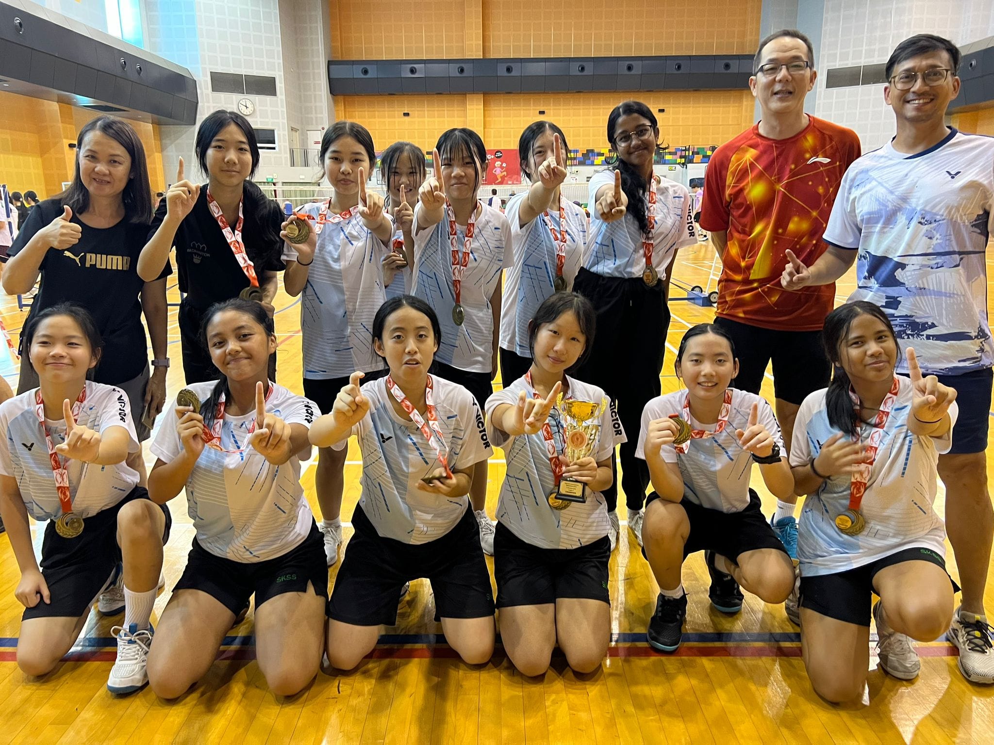 A group of young female athletes and coaches pose with medals and a trophy in a gymnasium, making victory signs.