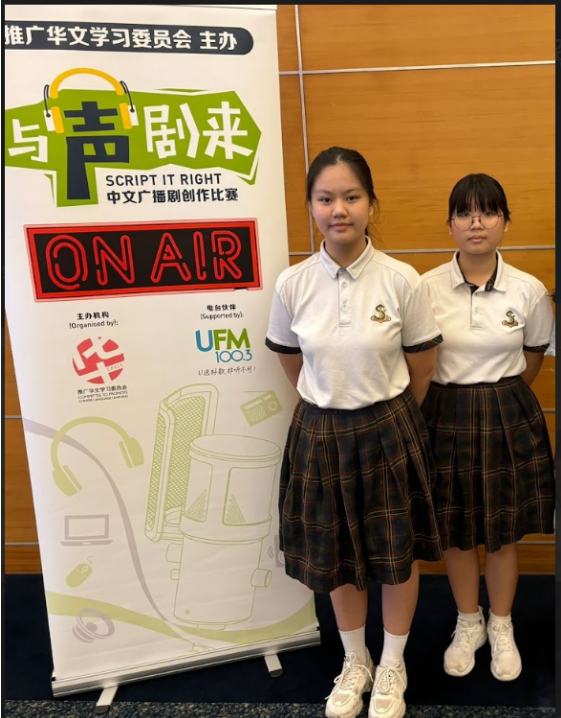 Two students in white school polo shirts and plaid skirts stand beside a banner for the “Script It Right” Chinese creative writing and radio drama competition. The banner features the text “ON AIR,” microphone graphics, and sponsor logos, positioned against a wood-paneled indoor wall.