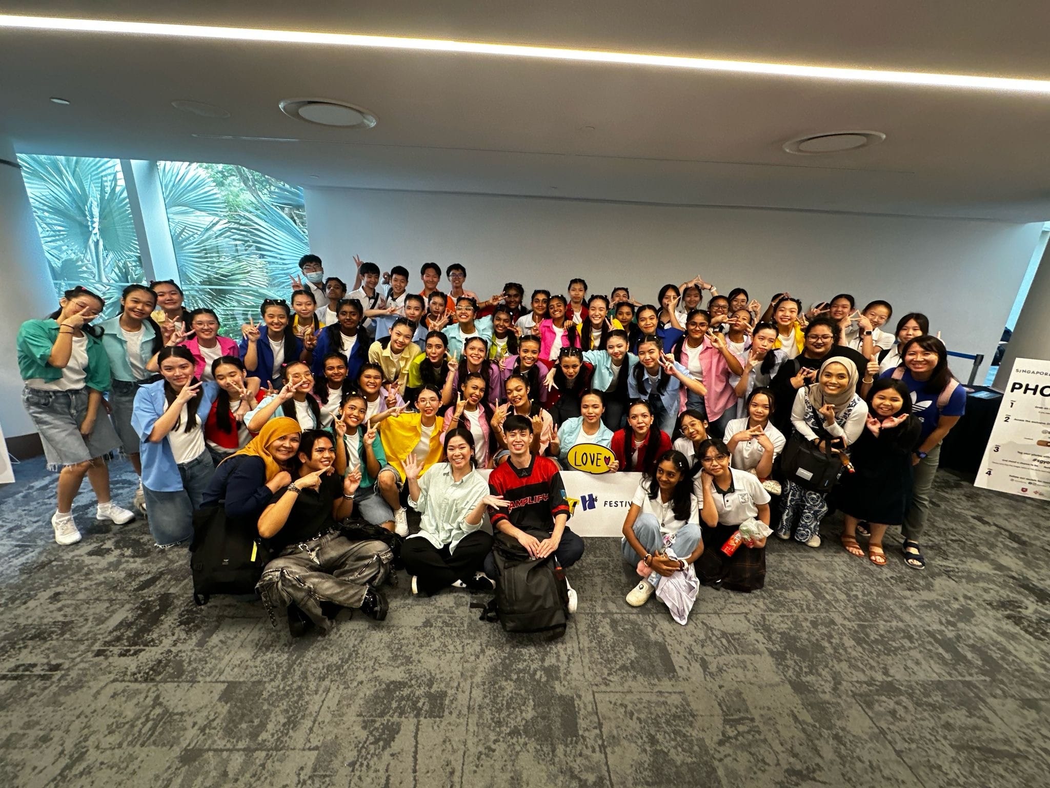 Large group of smiling young people posing indoors, many making peace signs, with a "LOVE" sign visible.