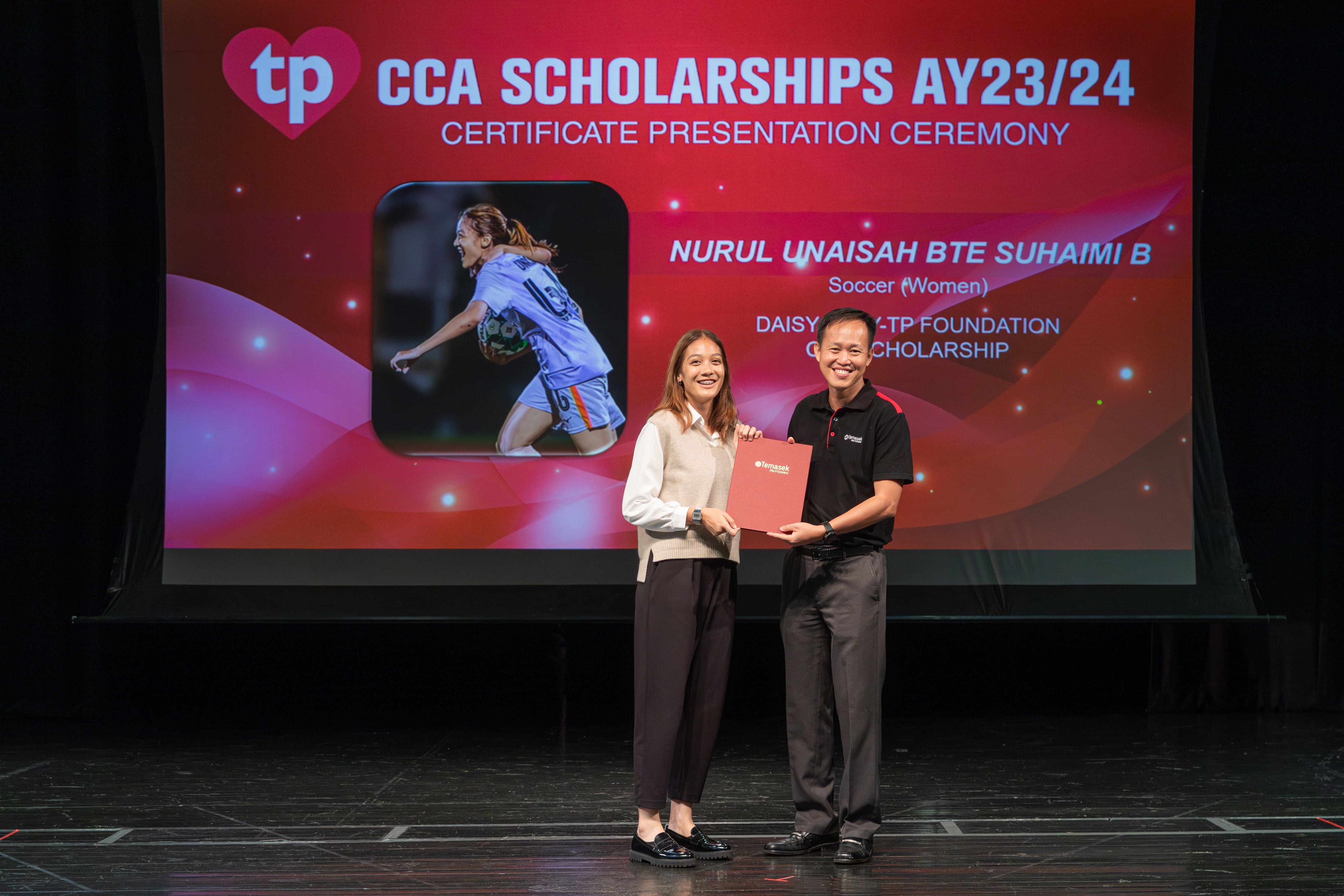 Two people on stage hold a red certificate folder before a screen reading CCA Scholarships AY23/24.