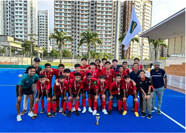 Boys hockey team in red jerseys posing with a trophy and medals on a blue turf field with buildings behind.