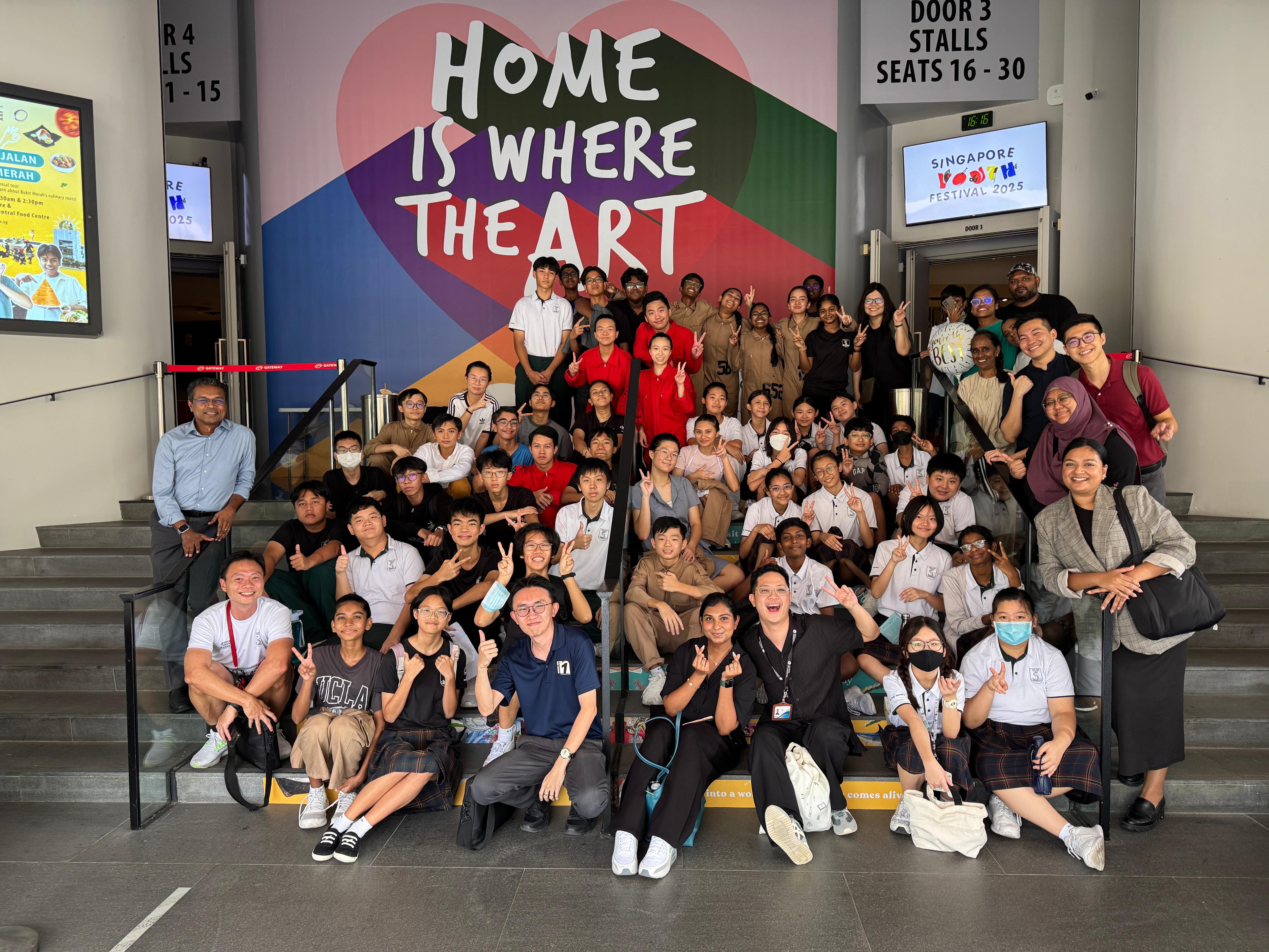Large group of students and adults posing on steps before a colorful mural reading "Home Is Where The Art."