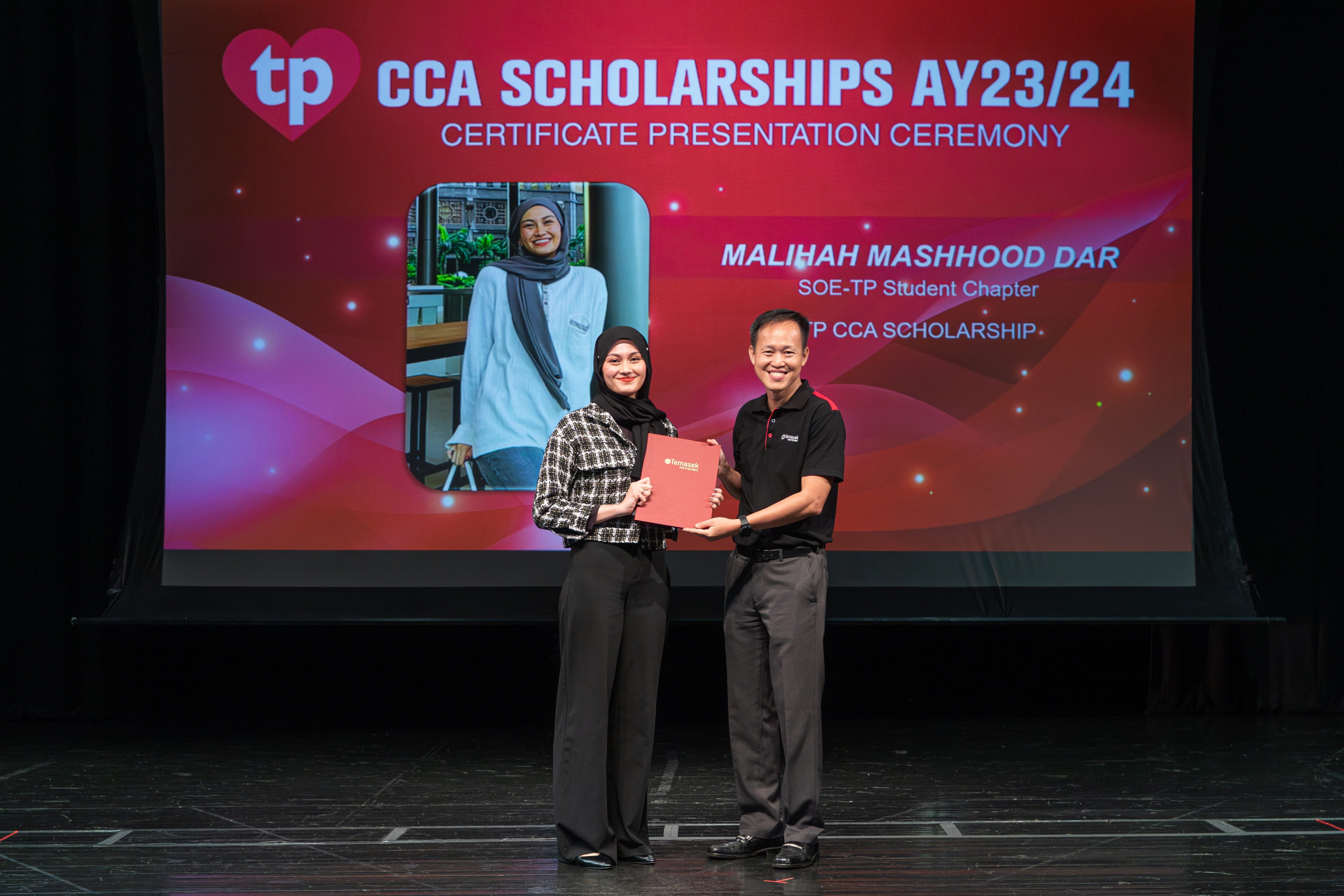 Two people hold a red certificate folder on stage at the TP CCA Scholarships AY23/24 Certificate Presentation Ceremony.