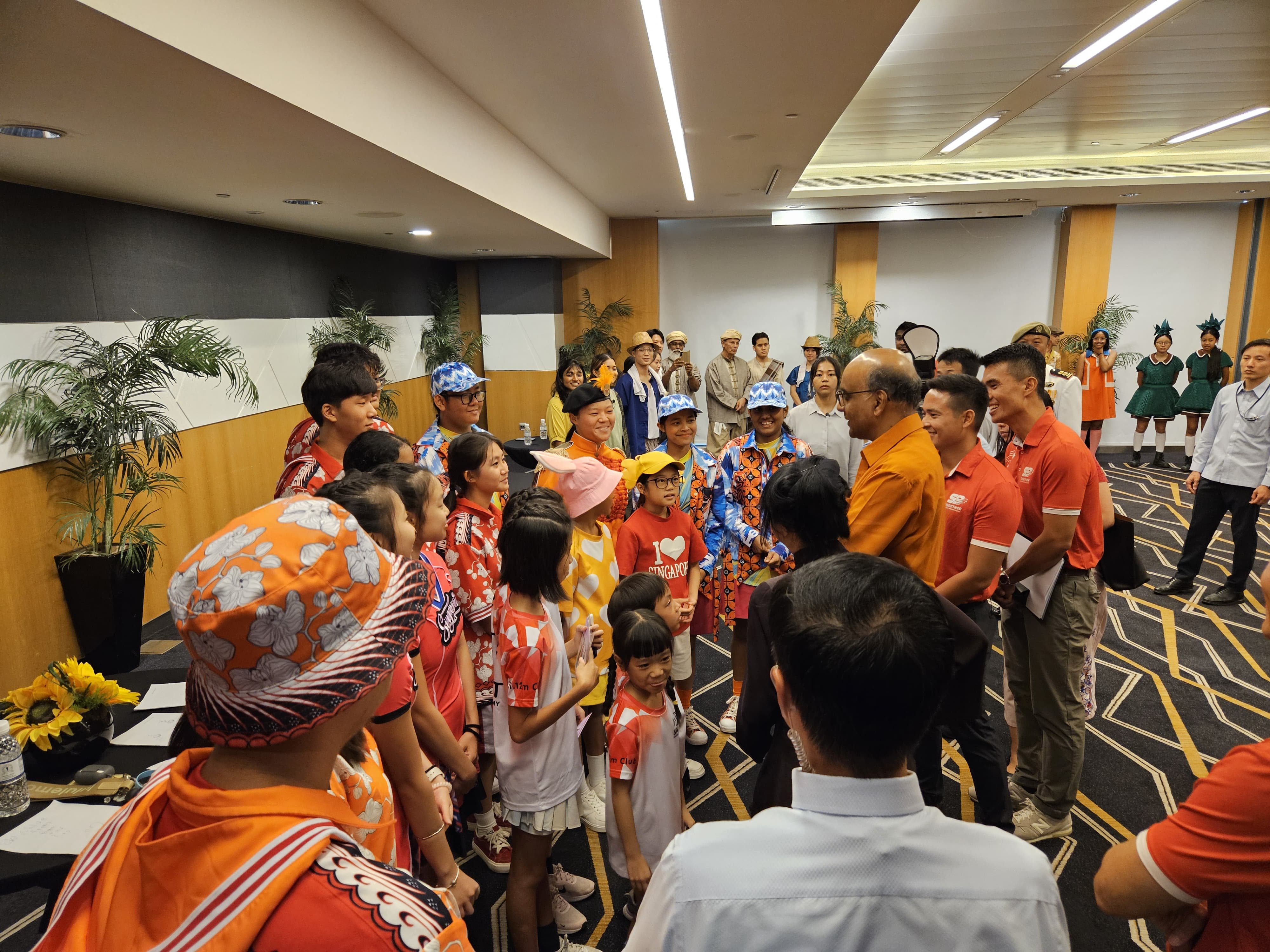 A large group of children and adults in colorful costumes gather indoors, with some wearing Singapore-themed shirts.