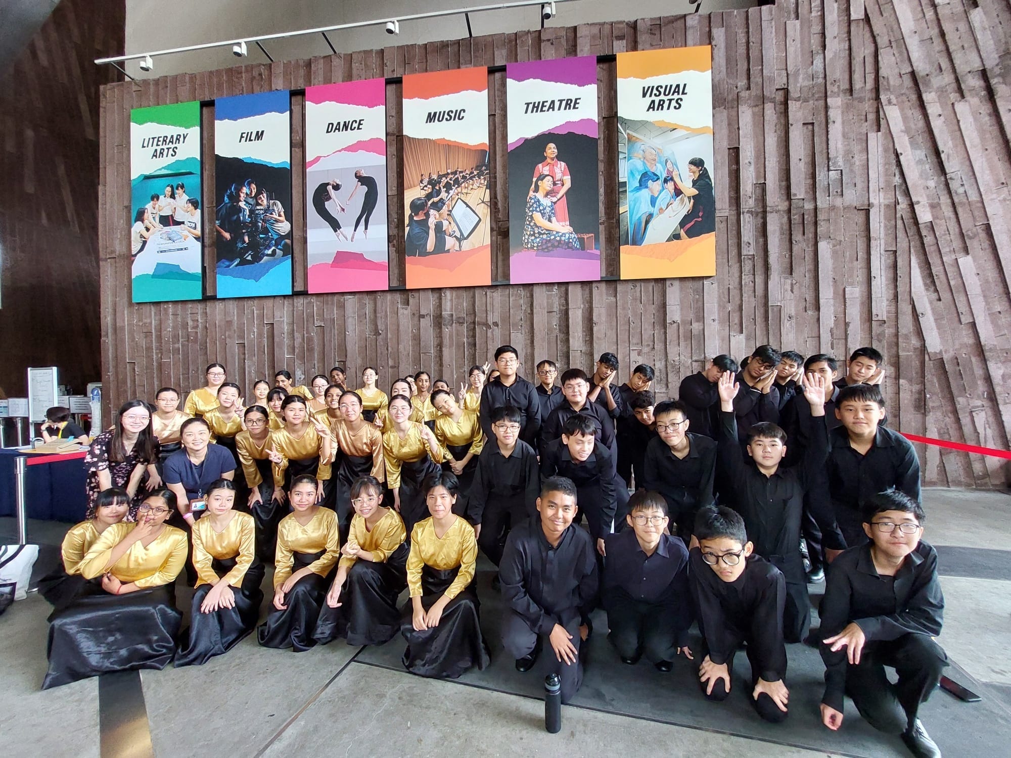 A large group of youth in gold and black outfits pose before colorful arts banners reading Film, Dance, Music, Theatre, and Visual Arts.