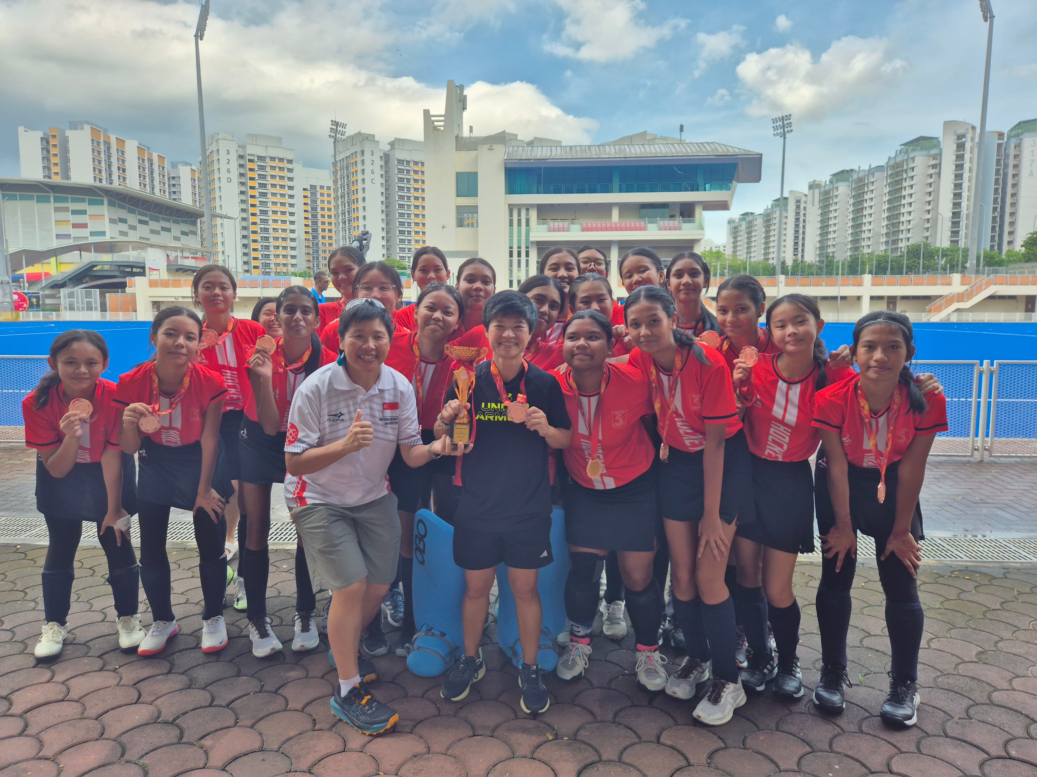 A large group of girls in red jerseys pose with medals and a trophy at an outdoor sports complex.
