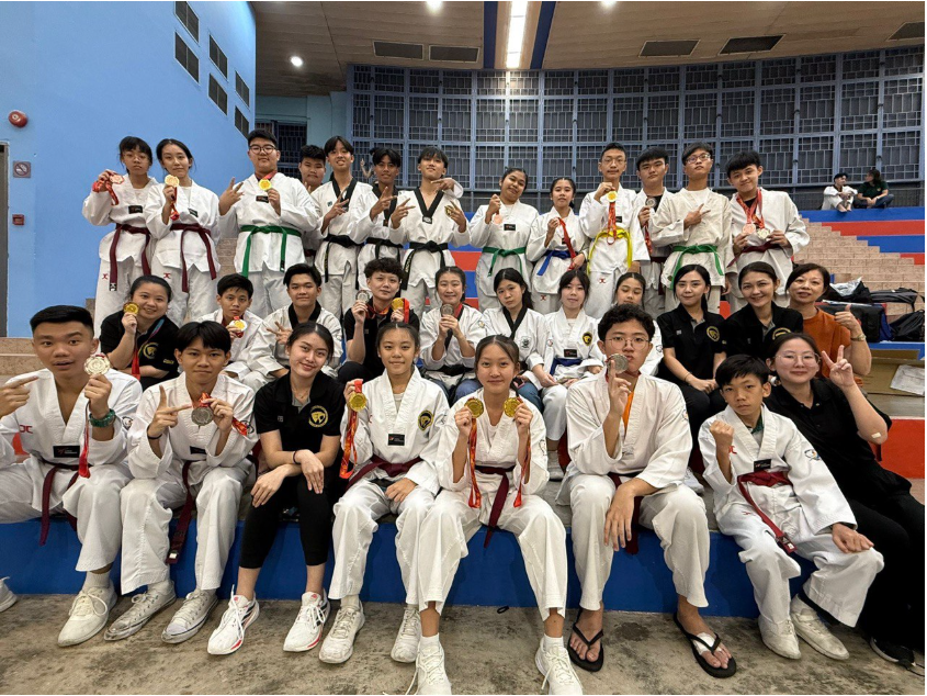 Large group of taekwondo students in white uniforms posing with gold medals in an indoor sports hall.