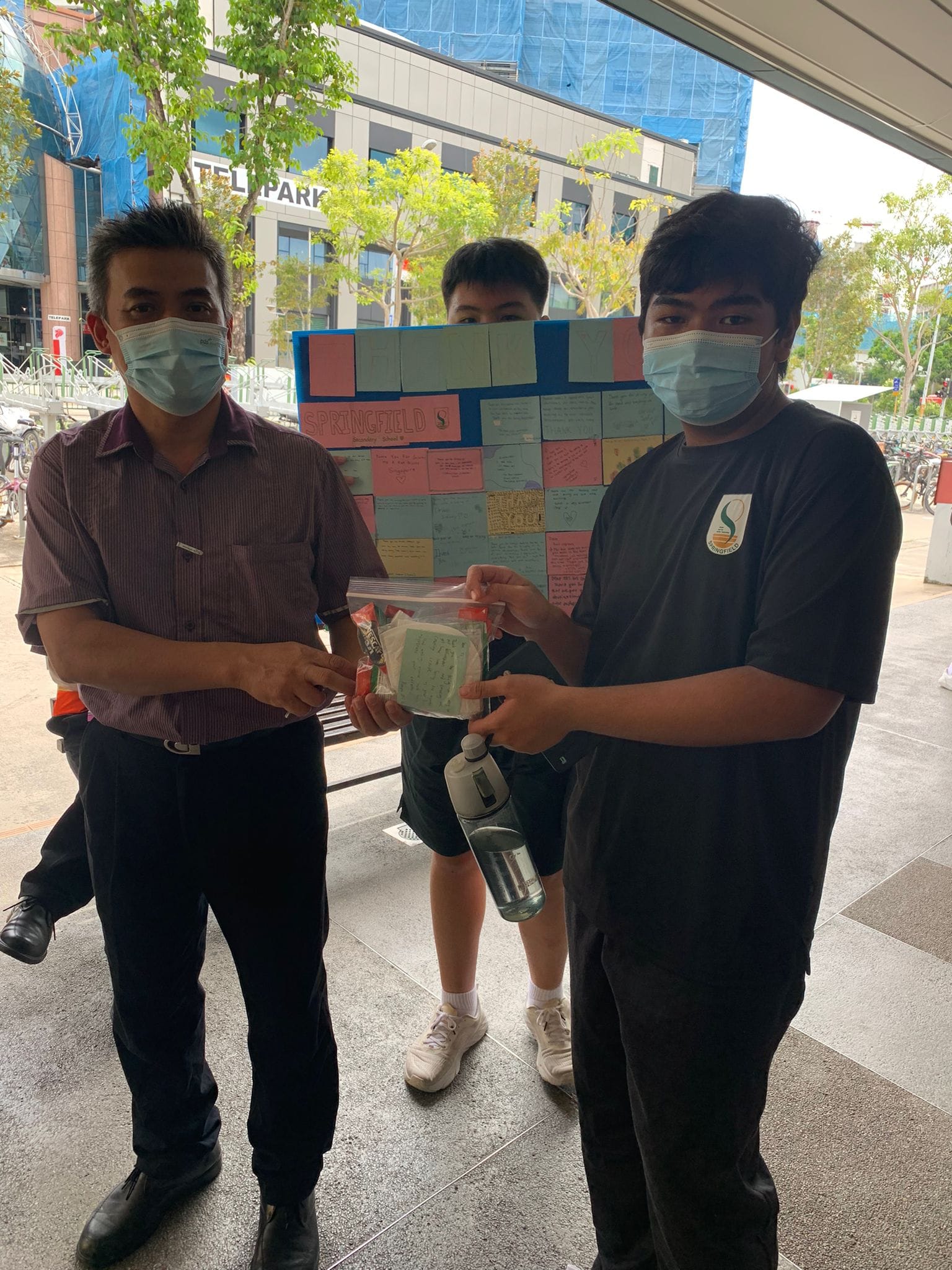 Three masked people outdoors holding a bag, with a colorful note-covered Springfield School board behind them.