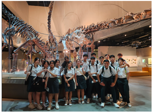Students in white shirts and dark skirts or pants pose in front of large dinosaur skeletons in a museum.