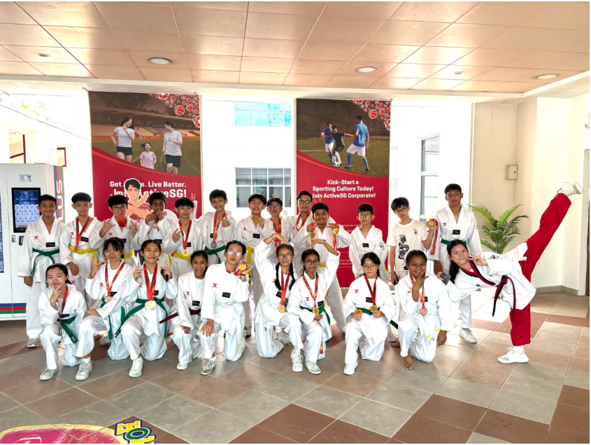 A group of young taekwondo students in white uniforms pose with medals in front of ActiveSG banners.