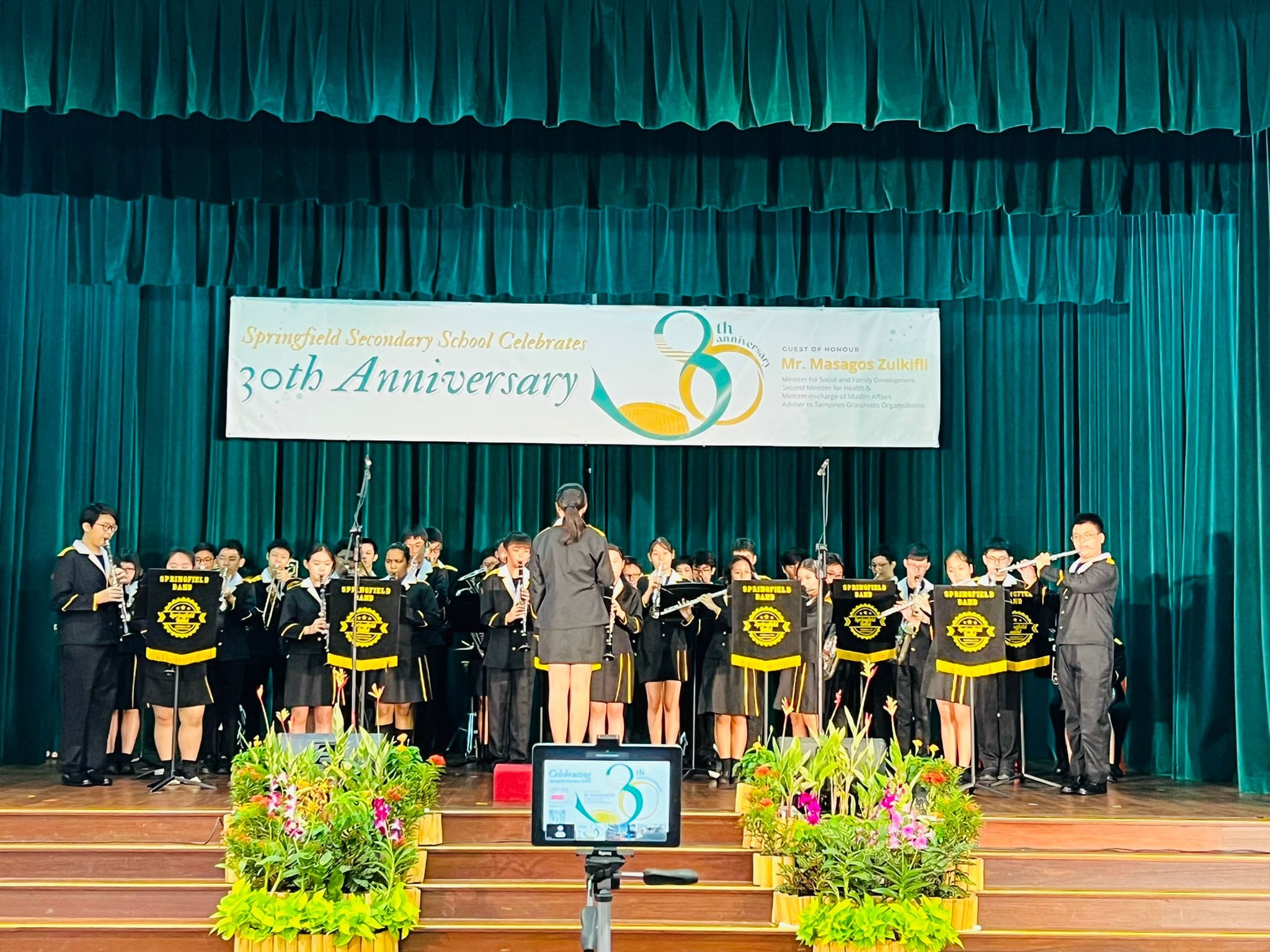 Springfield Secondary School band performs on stage at their 30th Anniversary celebration, with a conductor and uniformed students.