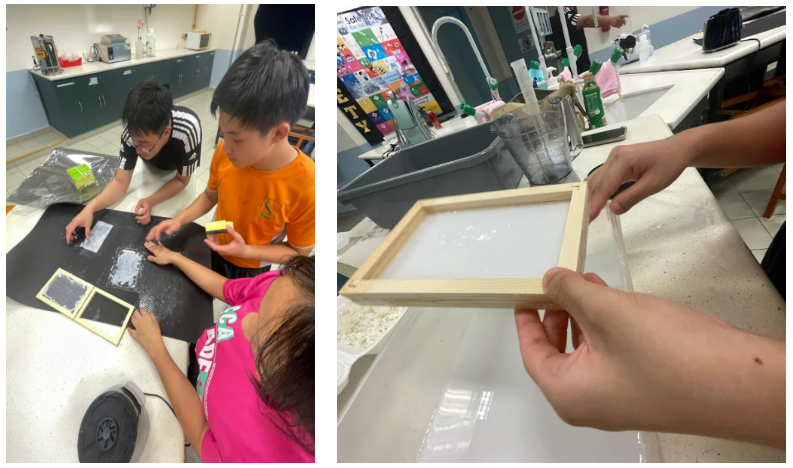 Three children work on a craft project at a dark table, while hands hold a wooden-framed screen nearby.