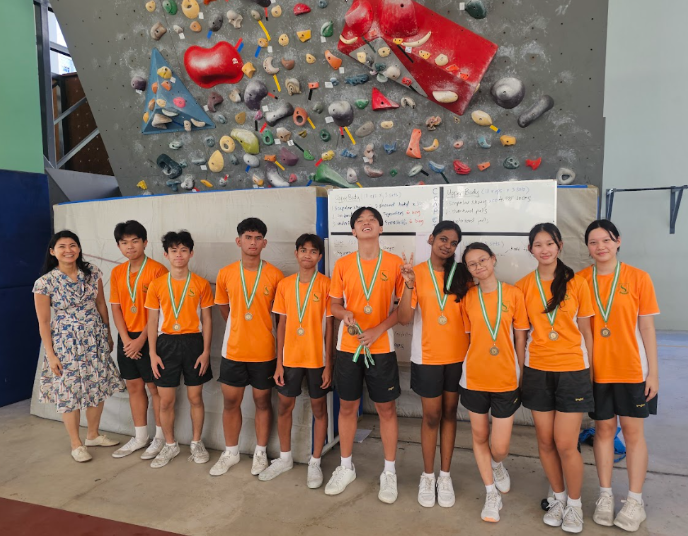 A group of students in orange shirts wearing medals stand in front of a colorful indoor climbing wall.