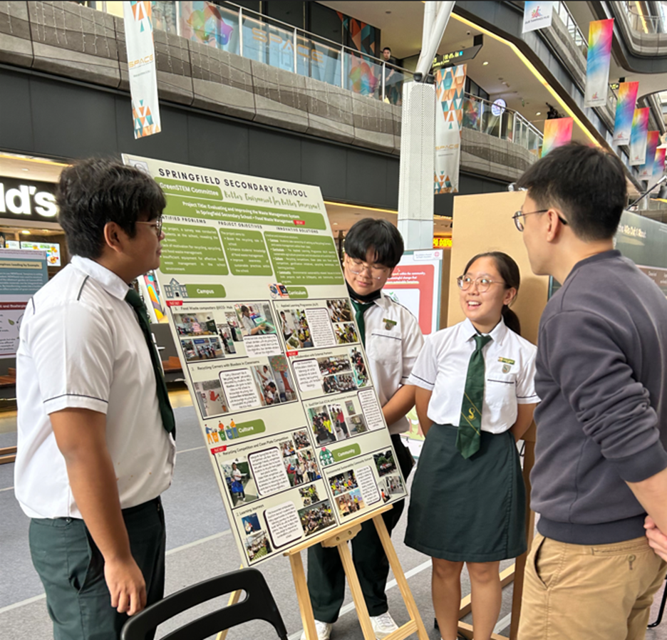 Three students in school uniforms present a Springfield Secondary School project poster to a visitor in a mall.