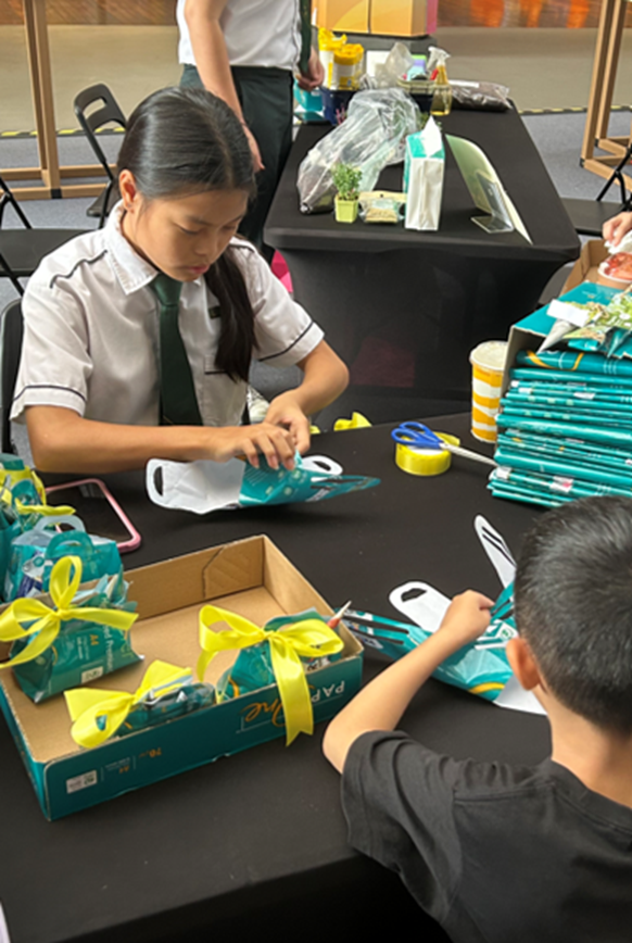Two children assemble teal gift bags with yellow ribbons at a black table with scissors and supplies.
