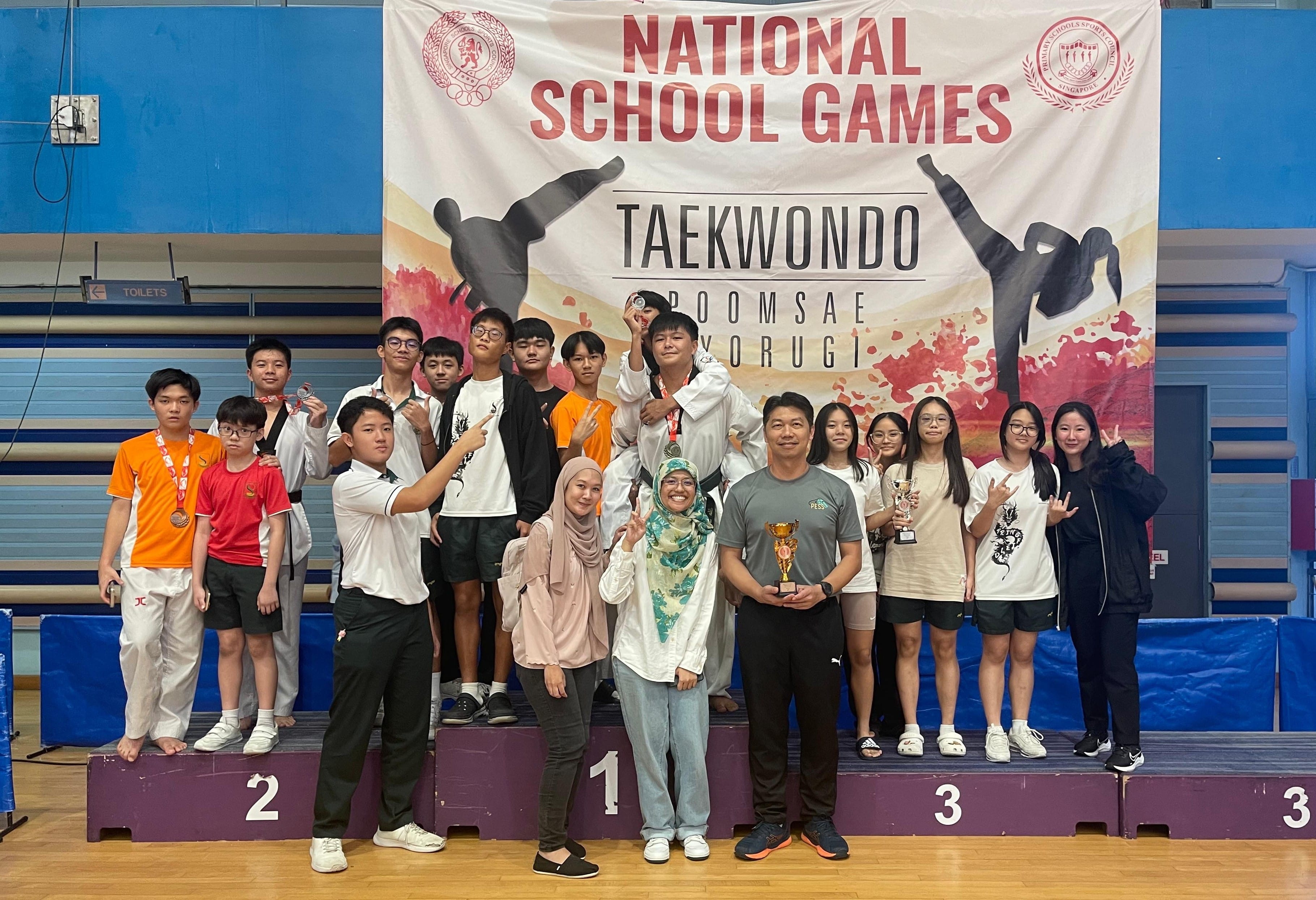 Group of students and coaches posing on a podium at the National School Games Taekwondo competition, holding trophies.