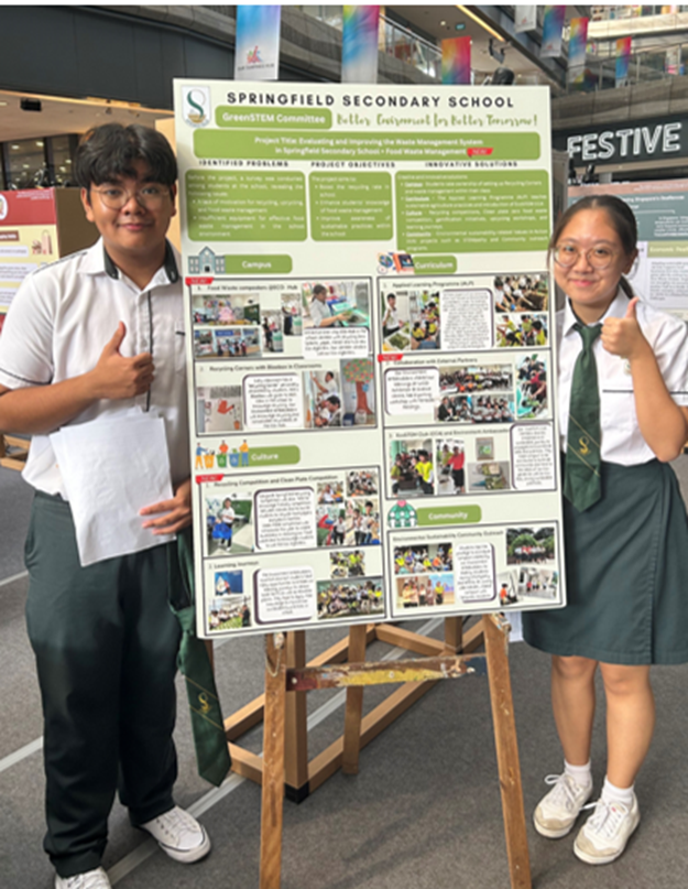 Two students in school uniforms give thumbs up beside a Springfield Secondary School GreenSTEM project poster on an easel.