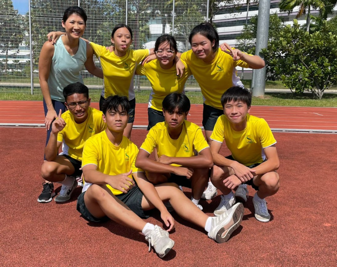 Eight students in yellow jerseys and an adult pose together on a red outdoor sports track.