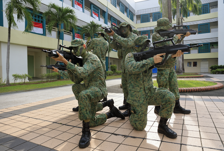 Four soldiers in green camouflage uniforms kneel and aim rifles in different directions outside a building.