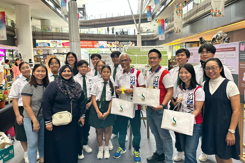 A group of students in green-and-white uniforms and adults pose together in a mall near a Springfield Secondary School display board.