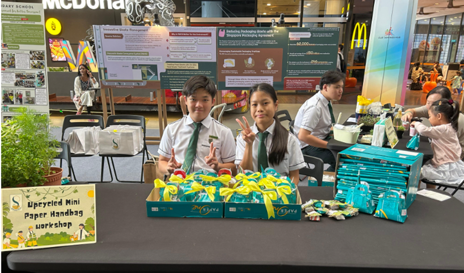Two students in uniforms pose at a table displaying an Upcycled Mini Paper Handbag Workshop booth in a mall.
