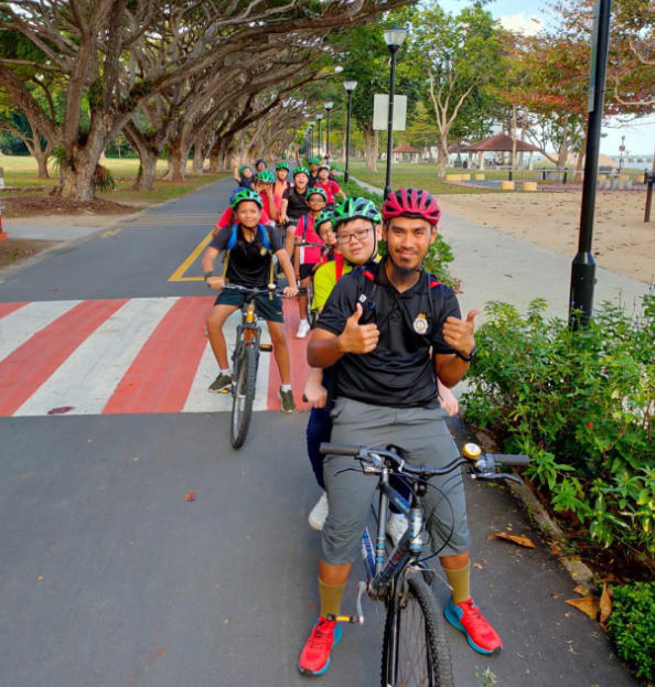 A group of children and an adult in helmets ride bicycles along a tree-lined park path, with the adult giving thumbs up.