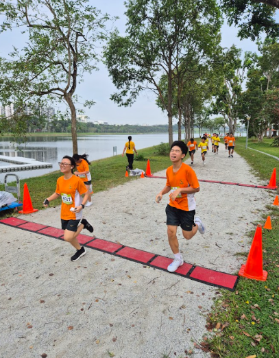 Runners in orange shirts cross a finish line on a park path beside a calm lake, with orange cones marking the route.
