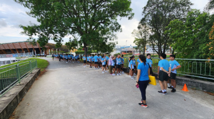 A long line of students in blue shirts walking along an outdoor path near trees and a building.