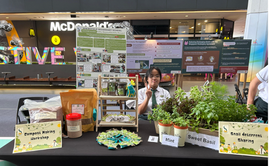 Student at Springfield Secondary School booth displaying mint, sweet basil, compost making workshop, and snail deterrent sharing.