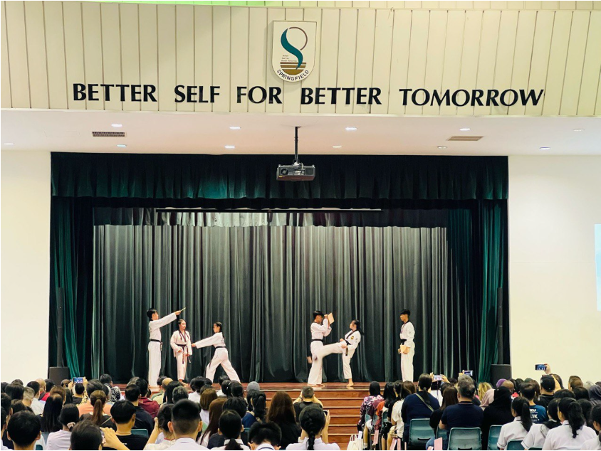 Six taekwondo students in white uniforms perform on a school stage before a seated audience.