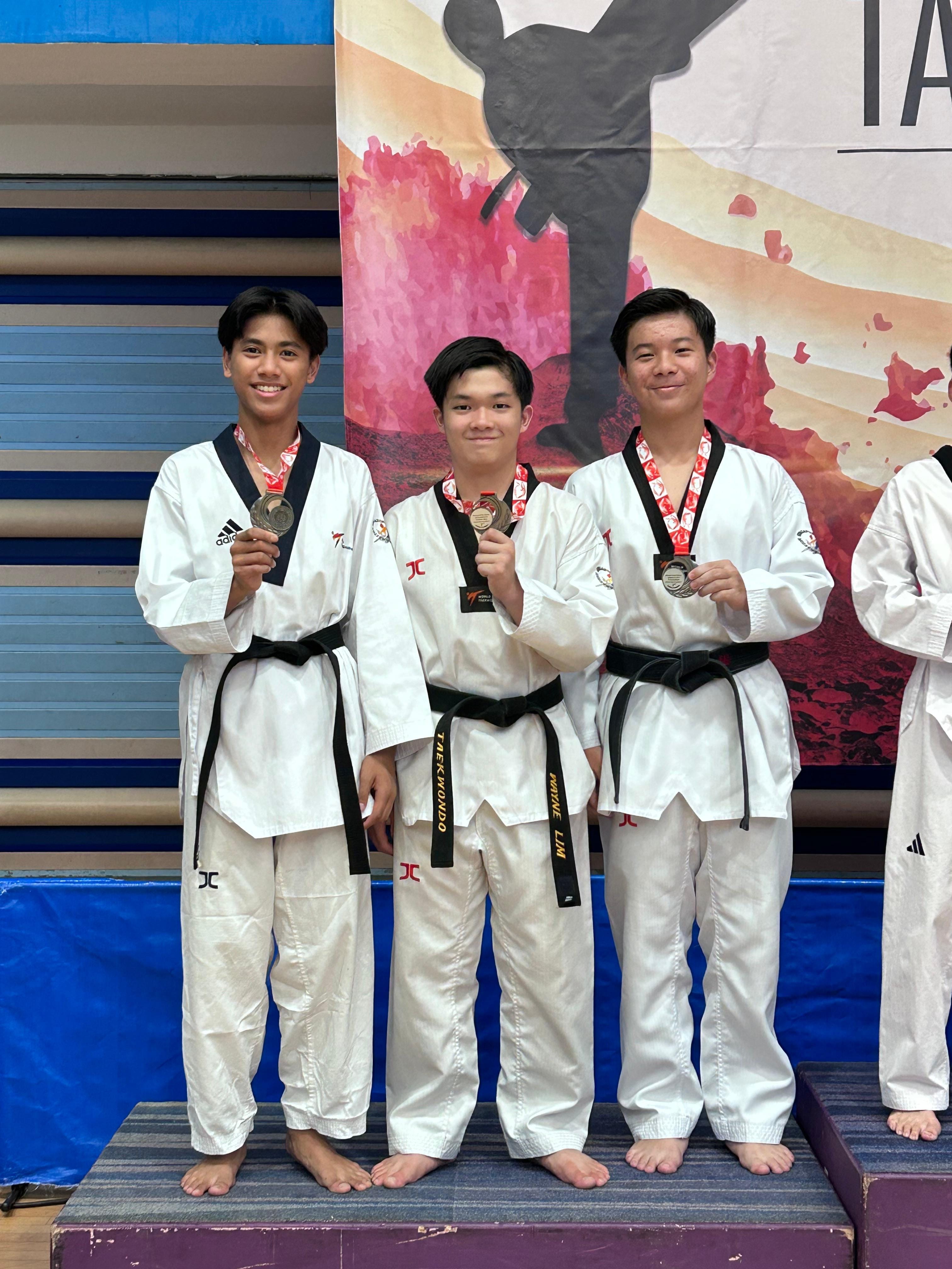 Three young taekwondo athletes in white uniforms with black belts hold up medals at a competition.