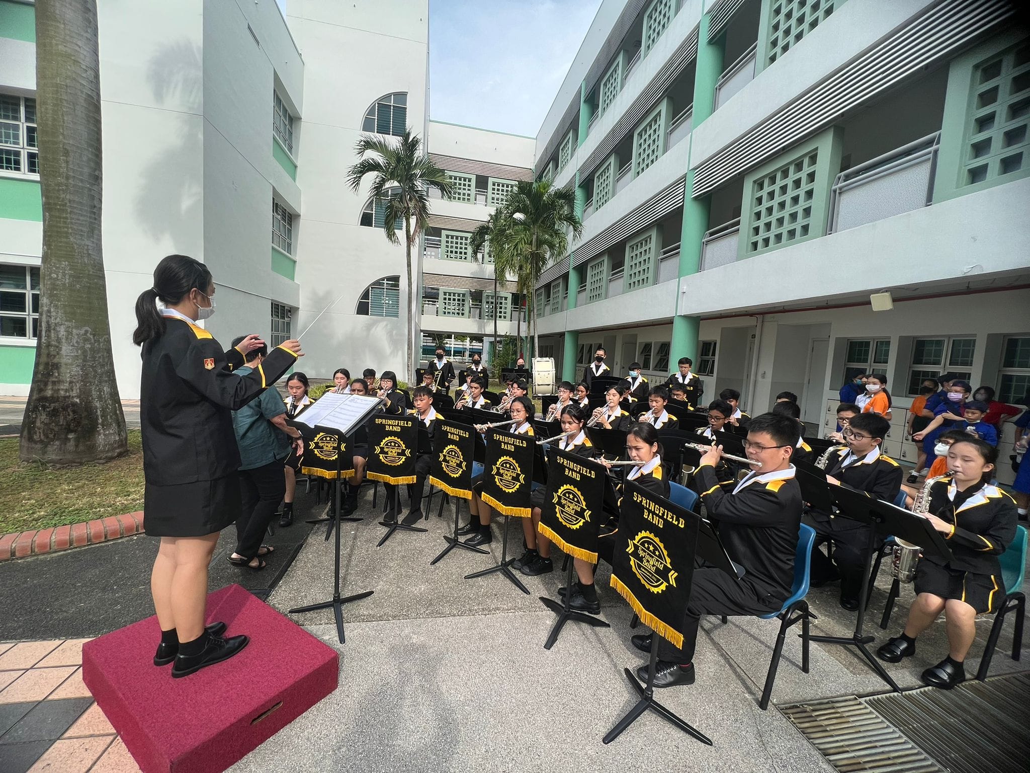 Student conductor leads the Springfield Band performing outdoors in a school courtyard, members in black uniforms playing wind instruments.
