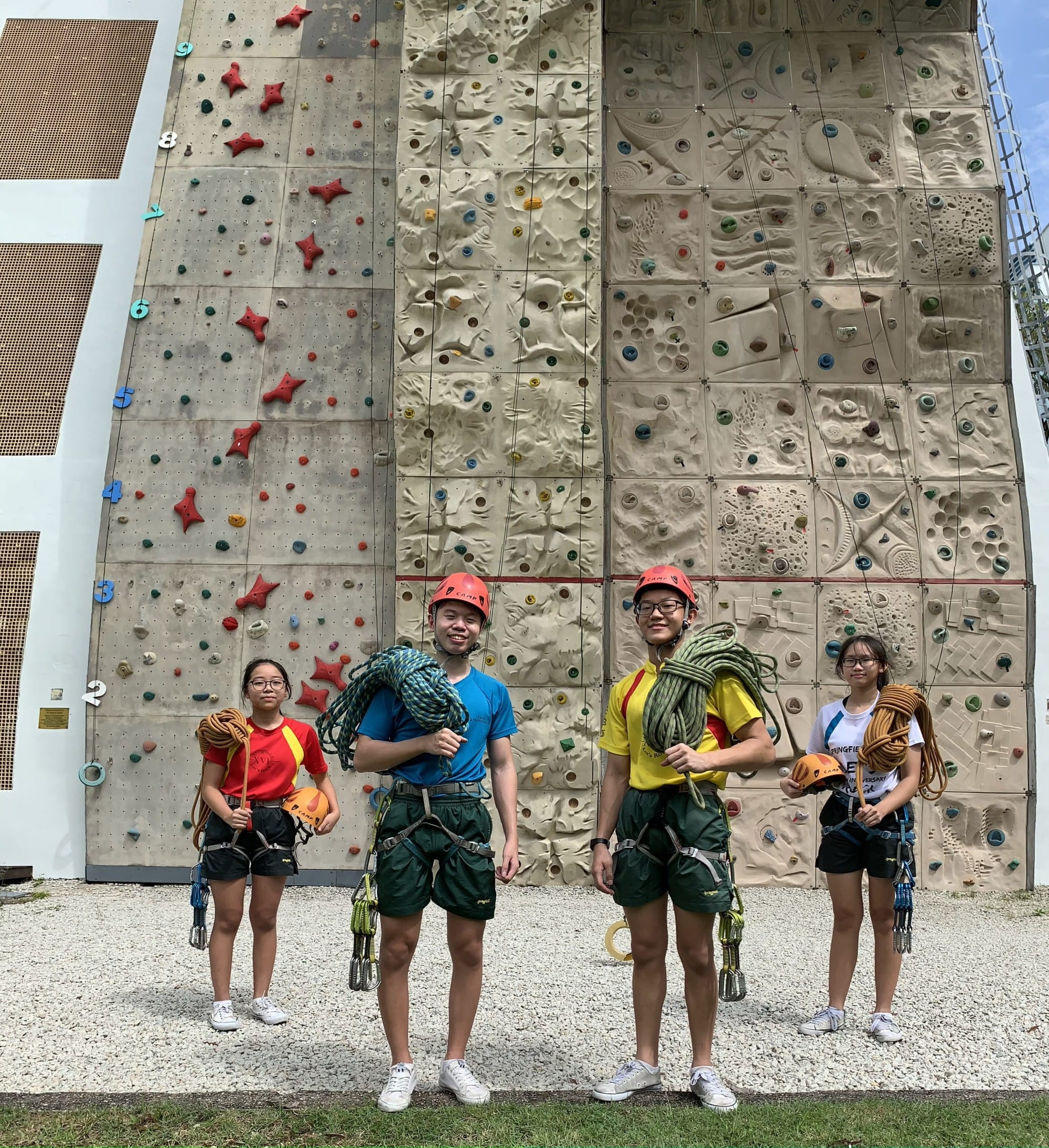 Four young people in harnesses and red helmets hold climbing ropes in front of a tall outdoor climbing wall.
