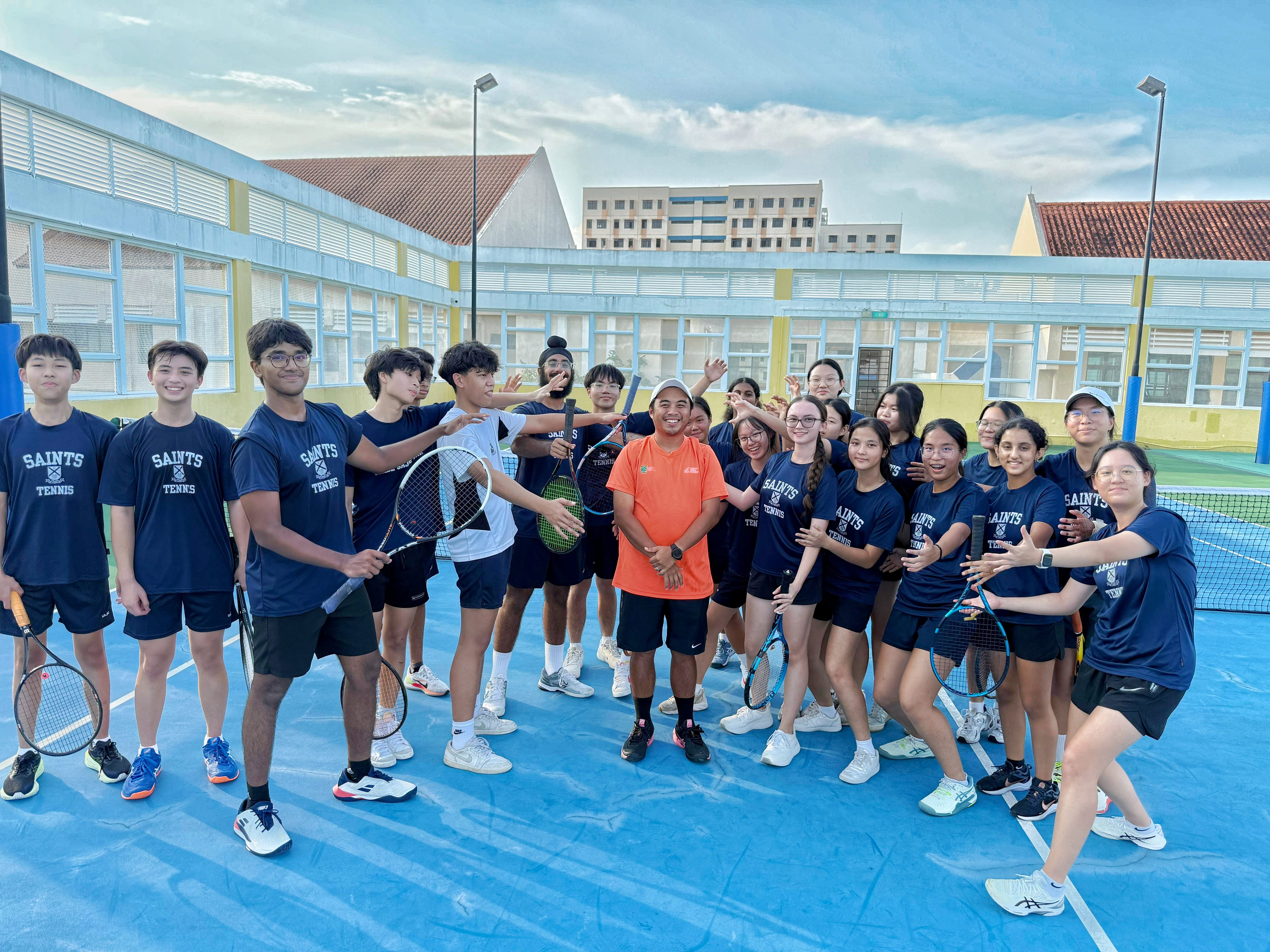 Saints Tennis players in navy blue 'Saints Tennis' t-shirts gather around a coach in an orange shirt on a blue outdoor tennis court, many holding tennis rackets and posing playfully for the photo.